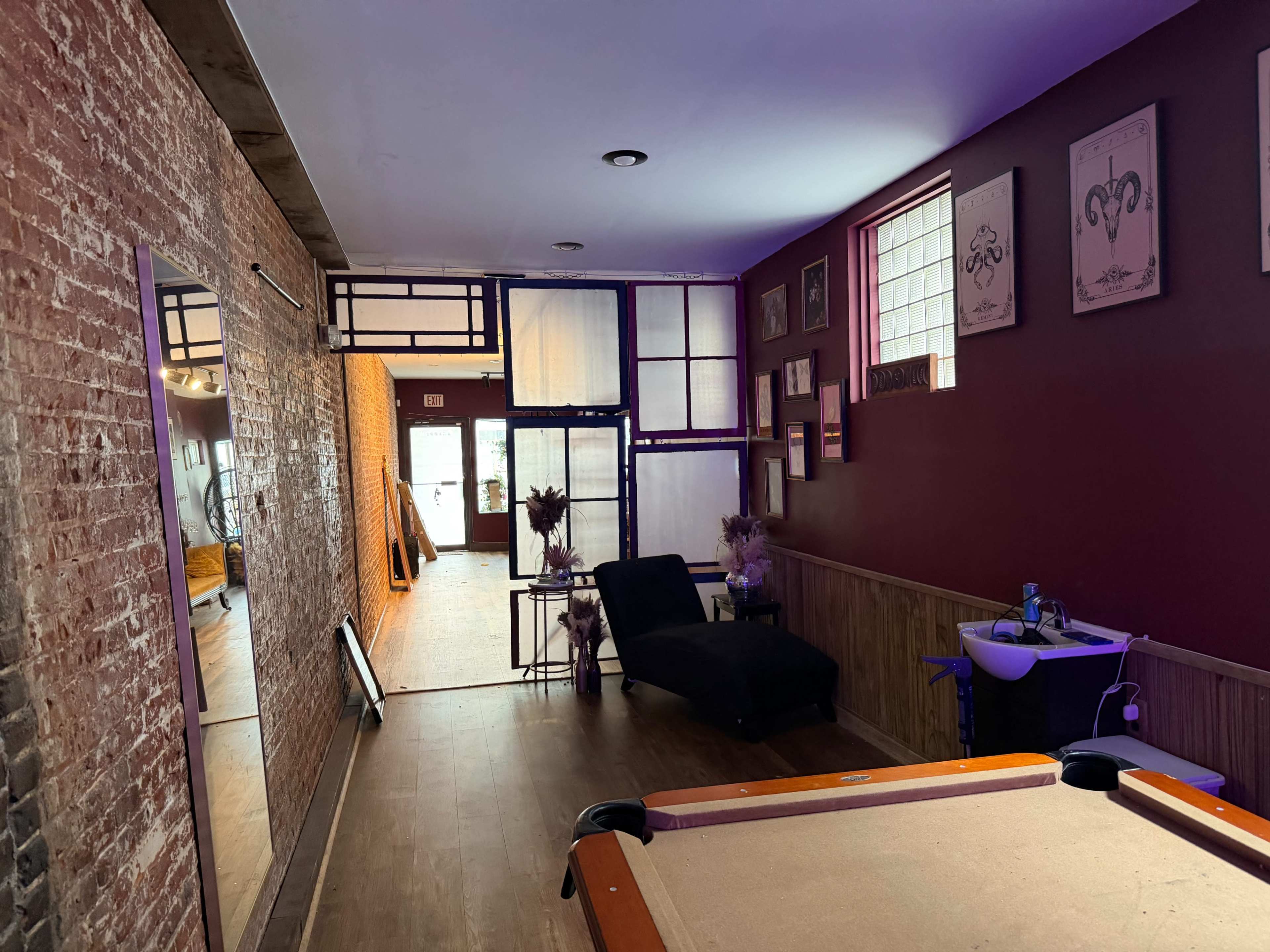 The image shows a room with a pool table, a long, narrow mirror on the wall, and a seating area, along with exposed brick walls and framed artwork.