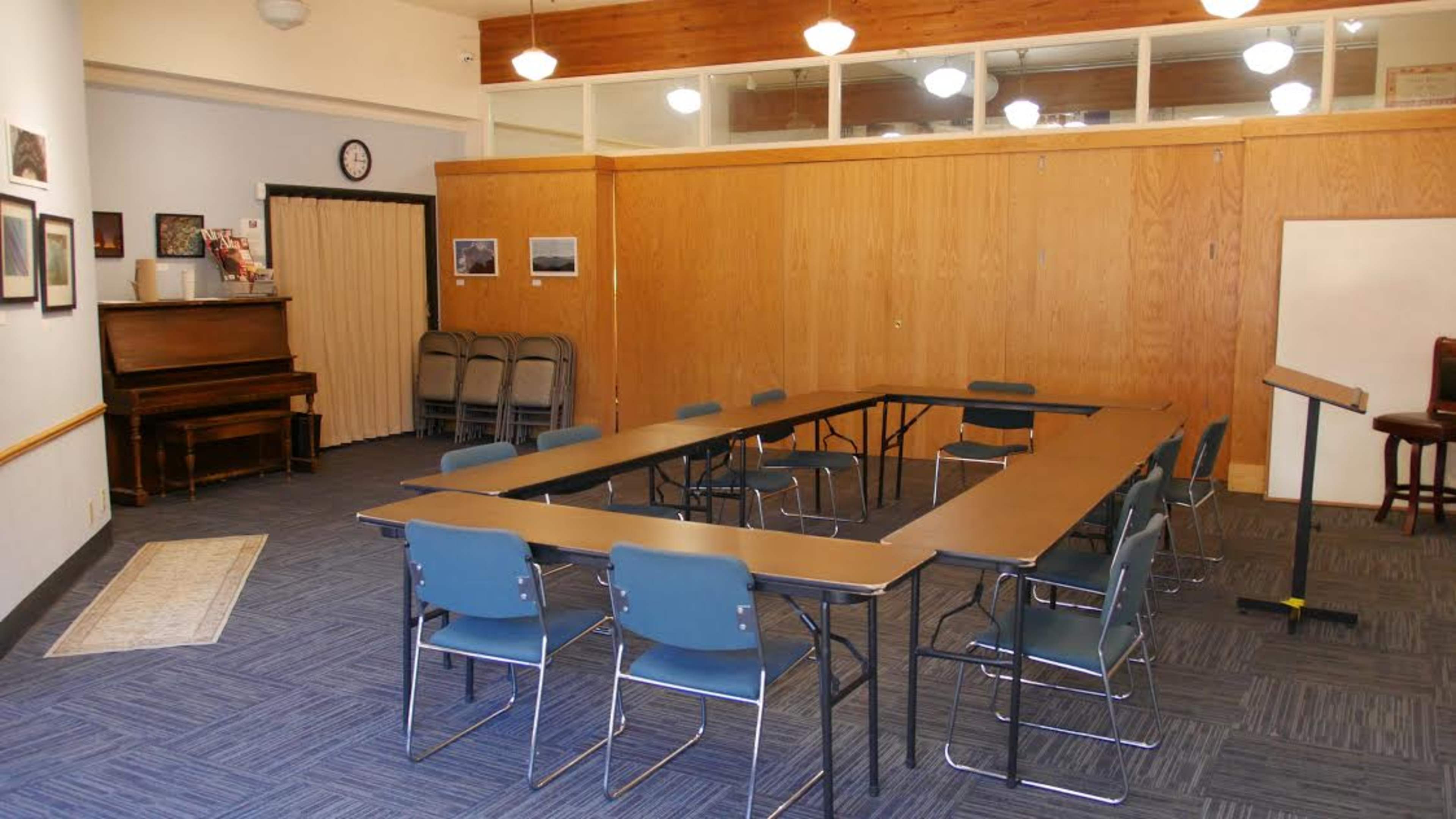 A large meeting room features an arrangement of tables and chairs in a rectangular setup, with a piano and additional seating against one wall.