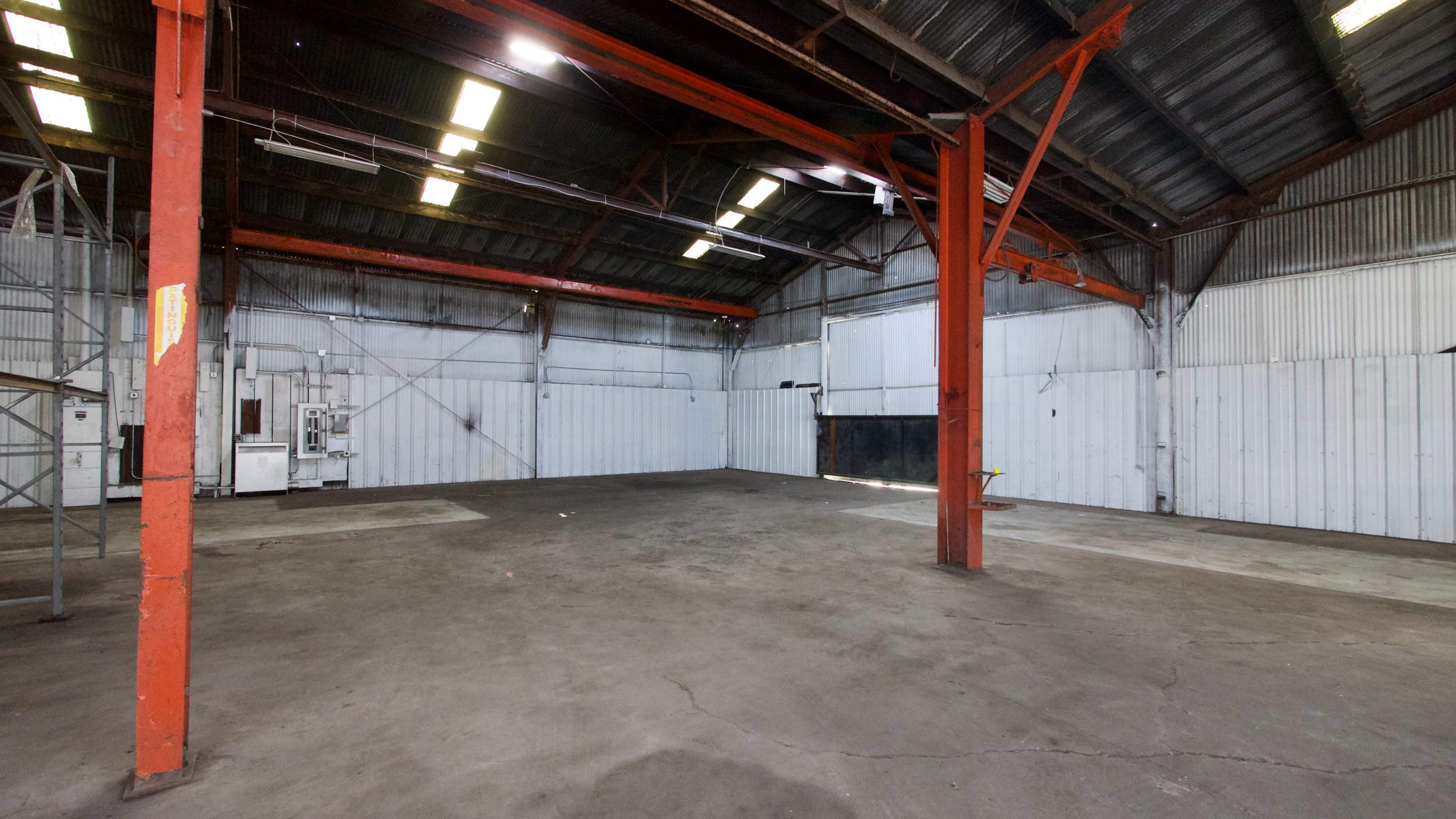 An empty warehouse features exposed metal beams, concrete flooring, and a high ceiling with fluorescent lights.