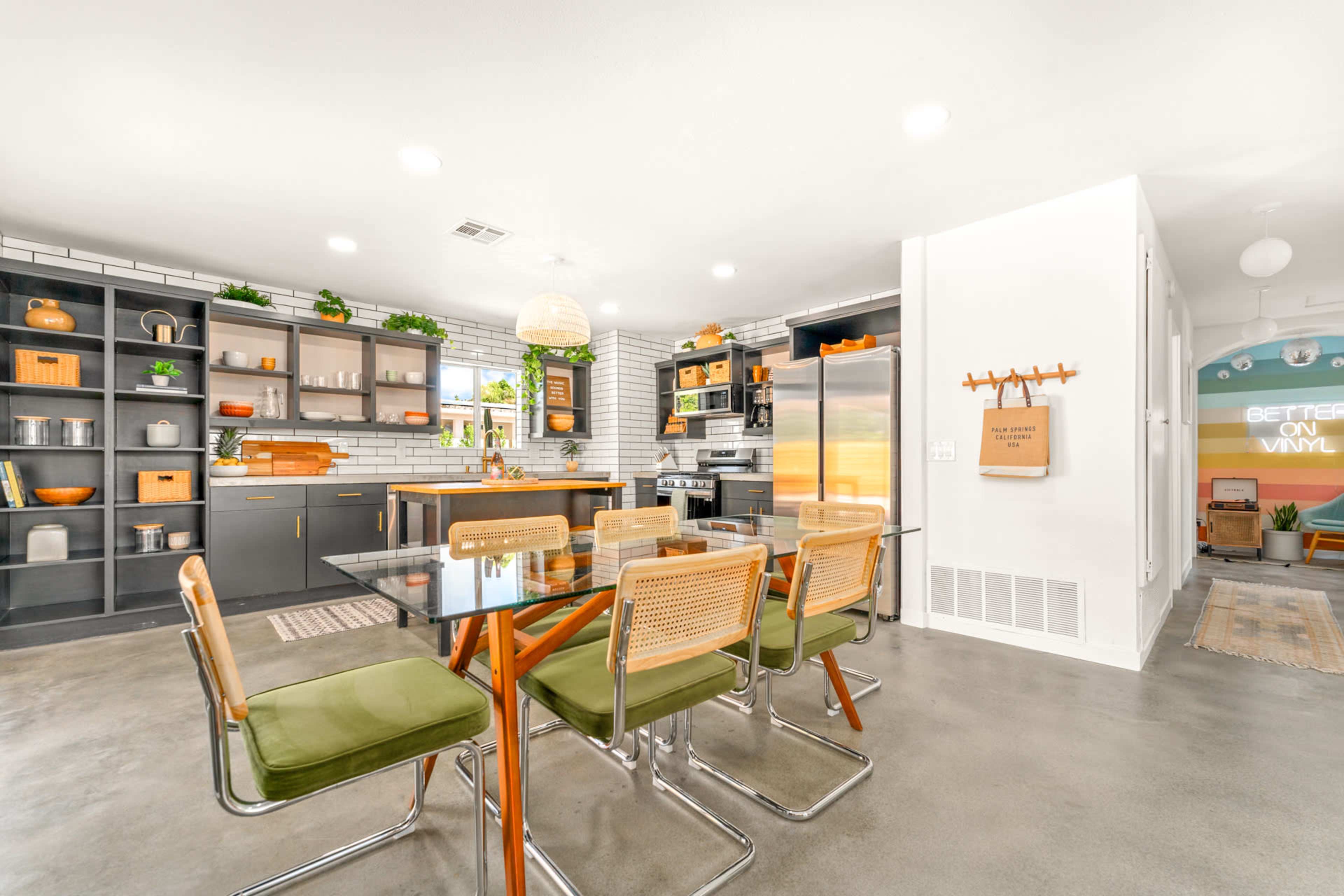A modern kitchen and dining area features a glass table with green chairs, alongside sleek cabinetry and stainless steel appliances.