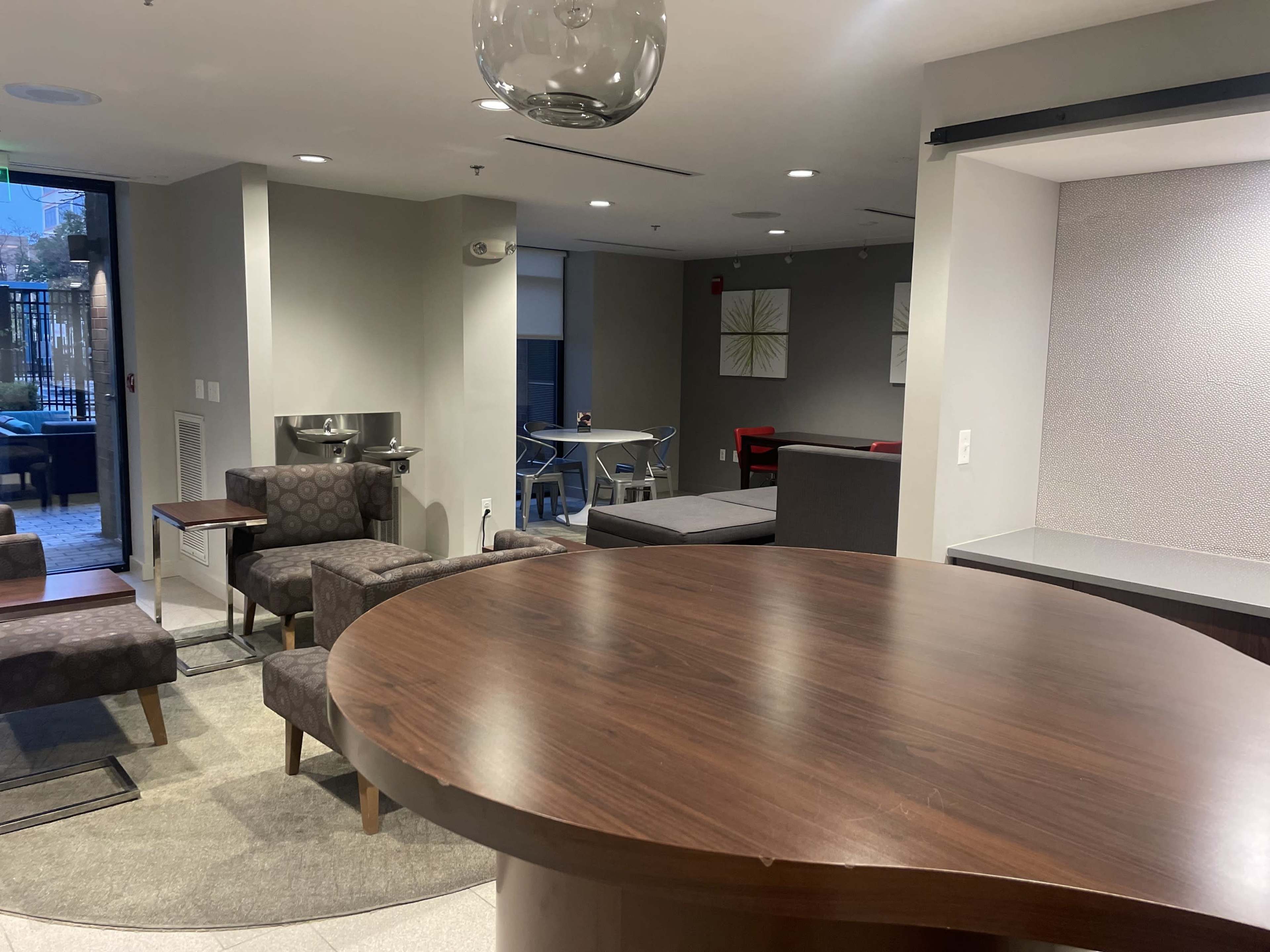 A modern lounge area with stylish seating, a circular wooden table, and a small kitchenette in a well-lit common space.