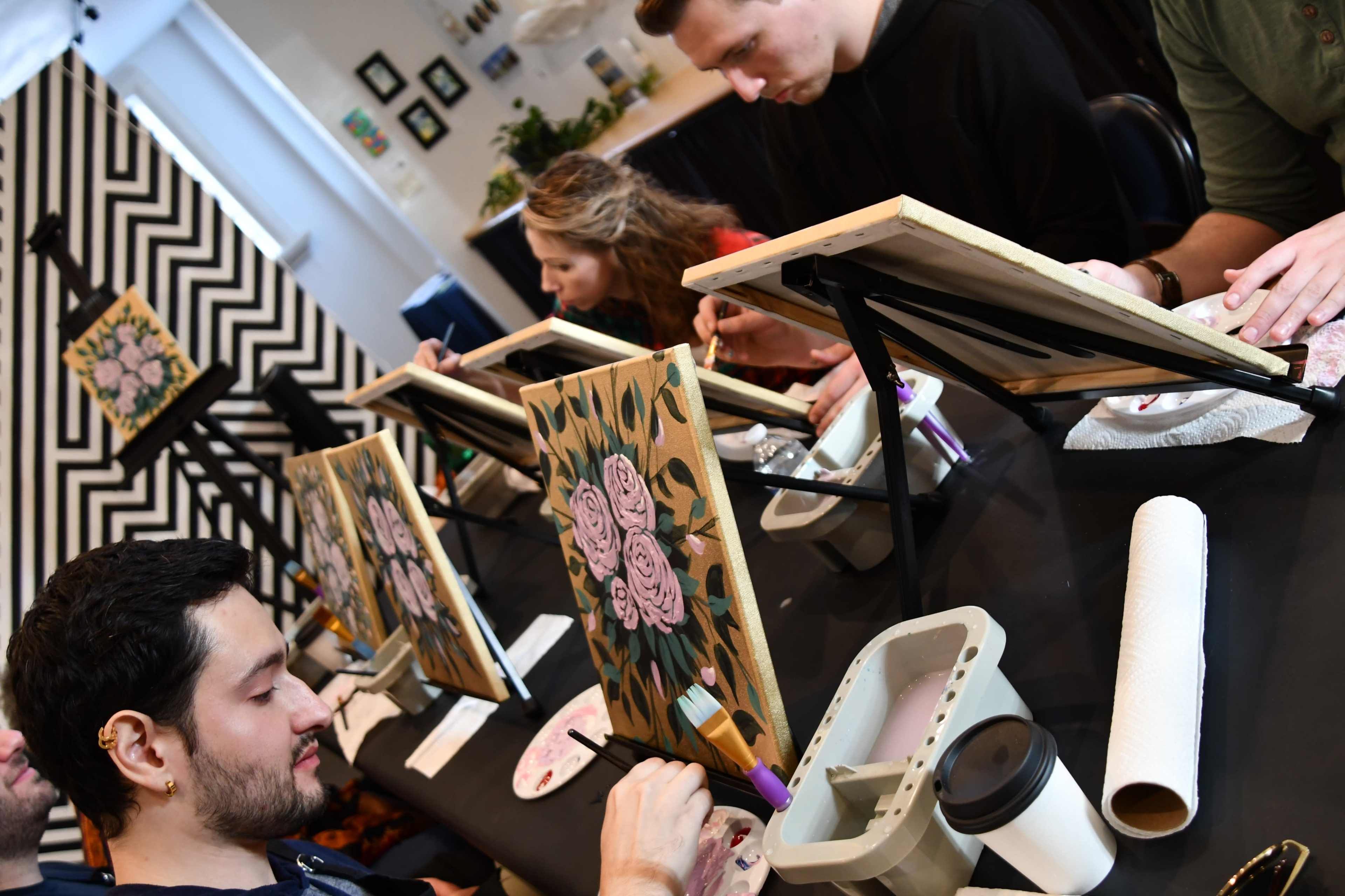A group of people are painting floral canvases at a table in a studio setting.