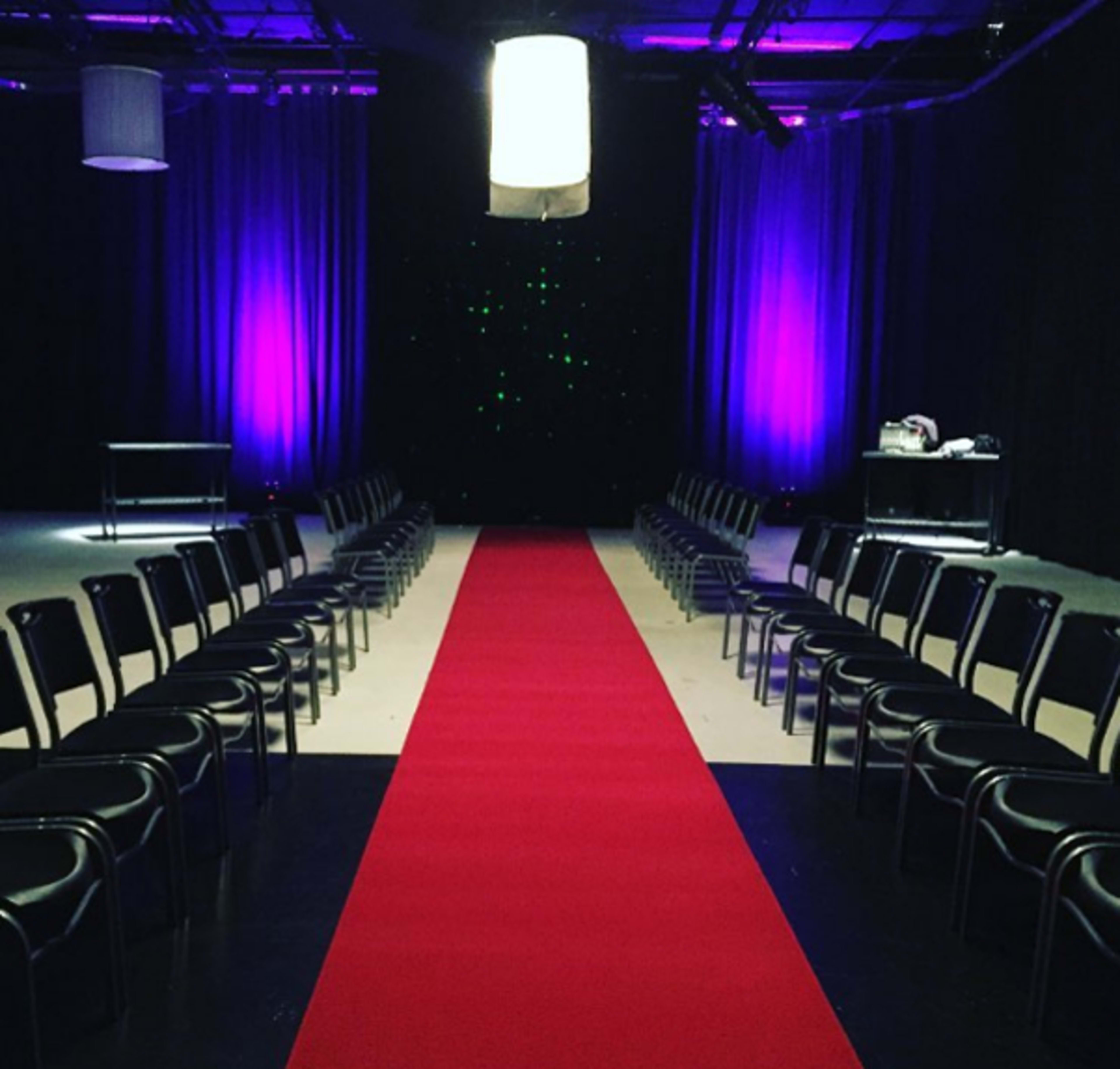 A red carpet runs down the center of a dimly lit room with rows of black chairs on either side and purple lighting in the background.
