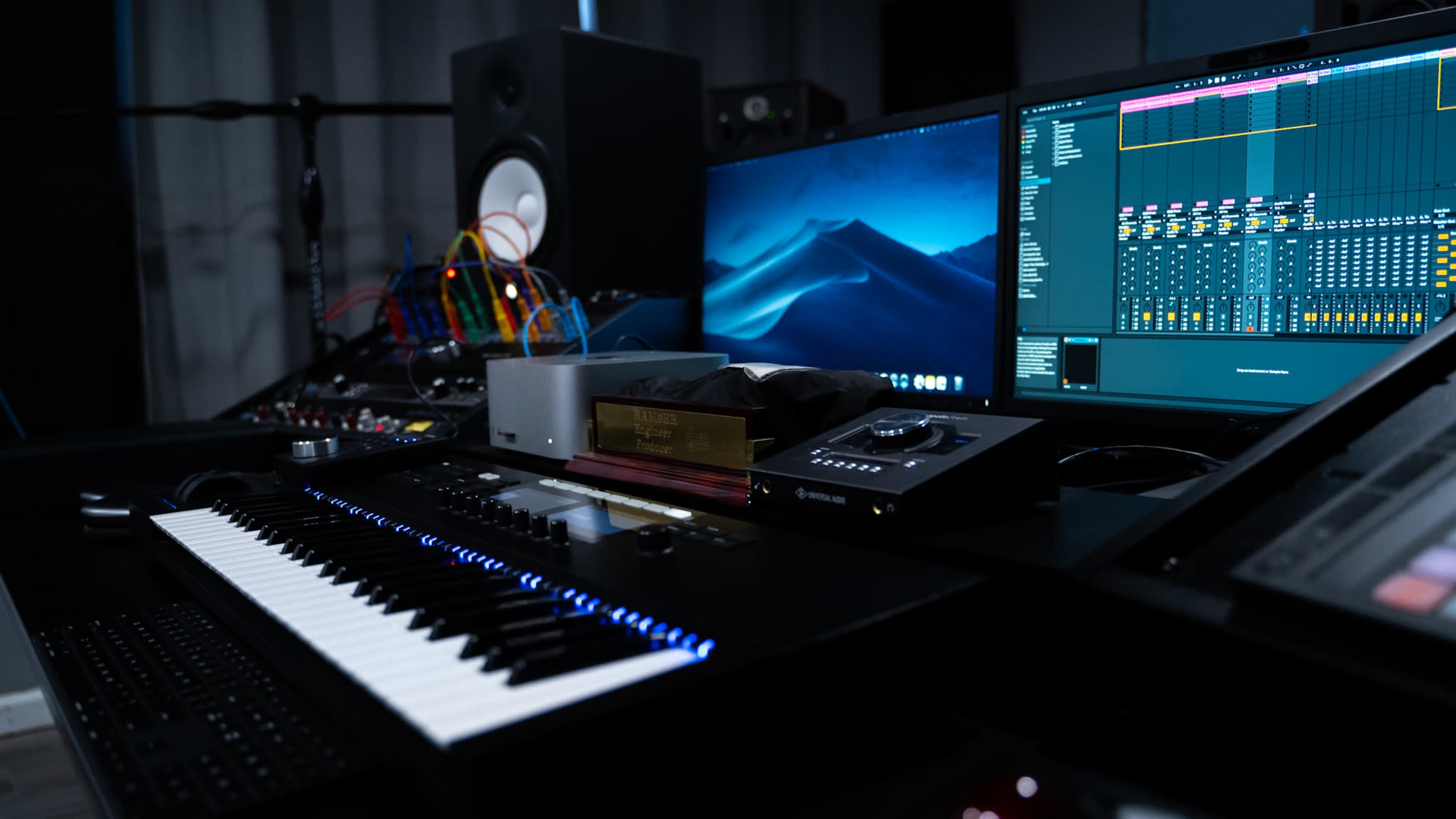A music production studio setup featuring a keyboard, computer screens with audio software, and various equipment including cables and a monitor speaker.
