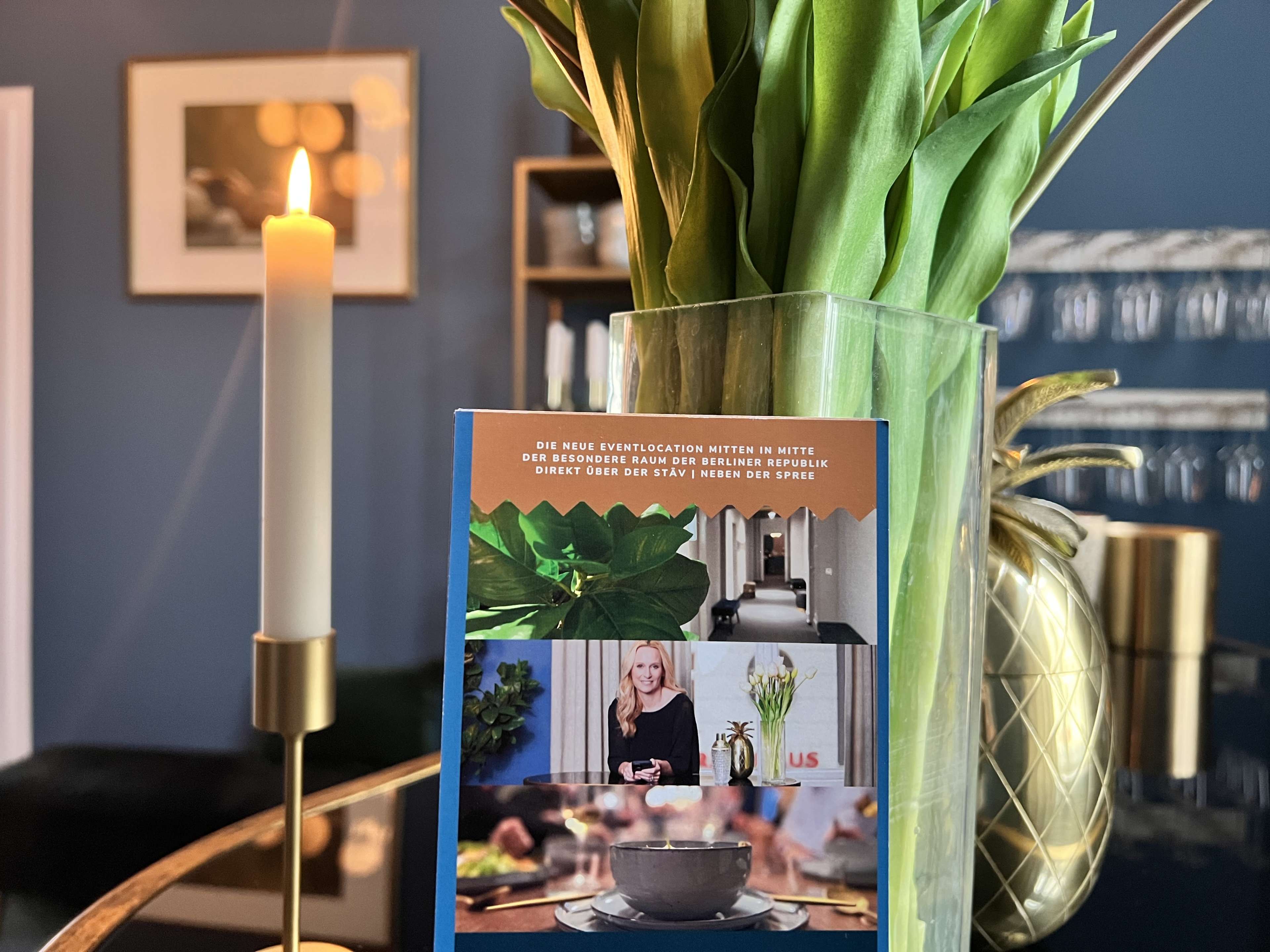 A decorative arrangement of fresh tulips is seen beside a lit candle and a postcard showcasing a restaurant interior and culinary imagery.