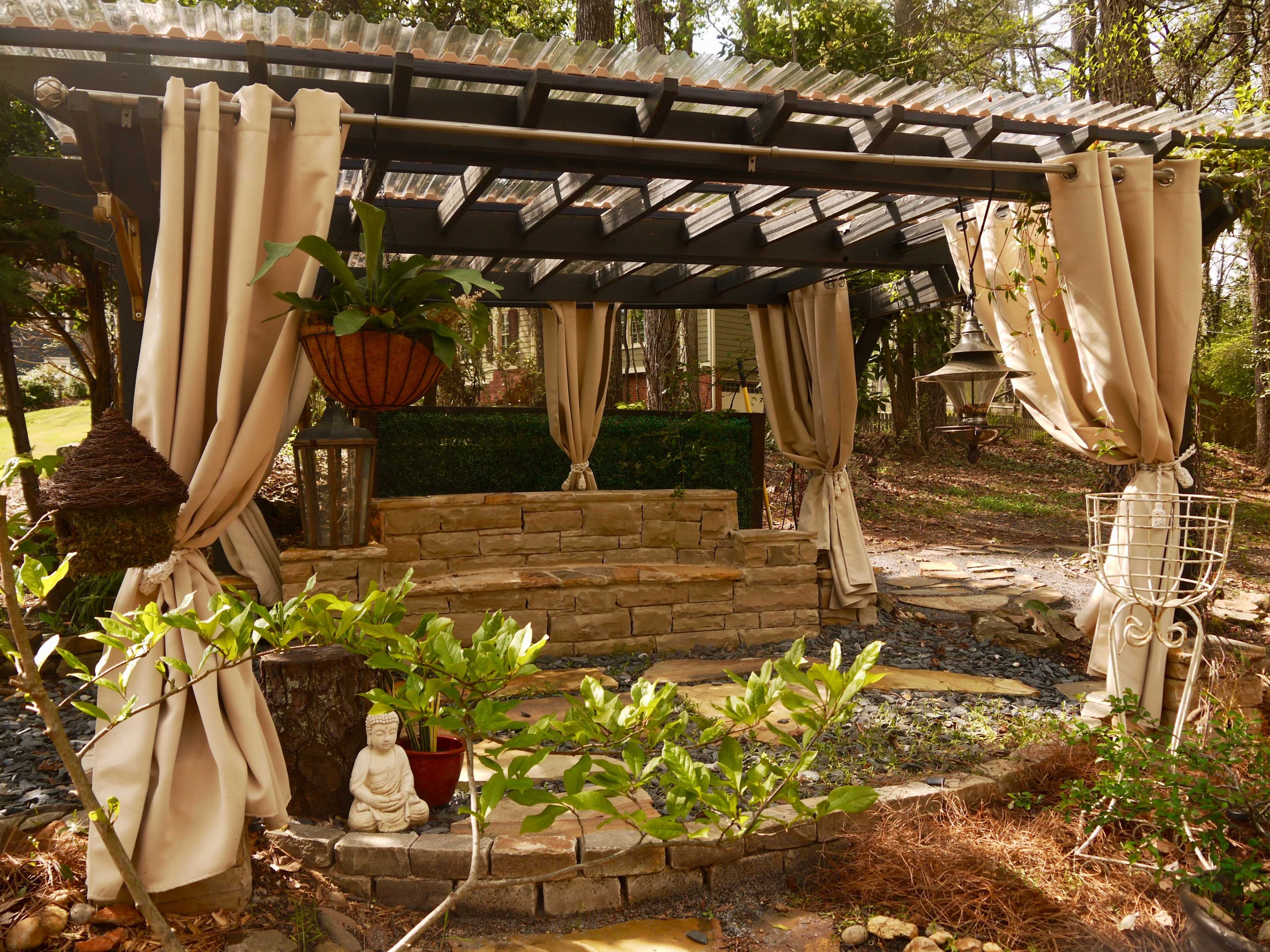 A gazebo-like structure with draped curtains, a stone seating area, and a small statue in a landscaped garden.