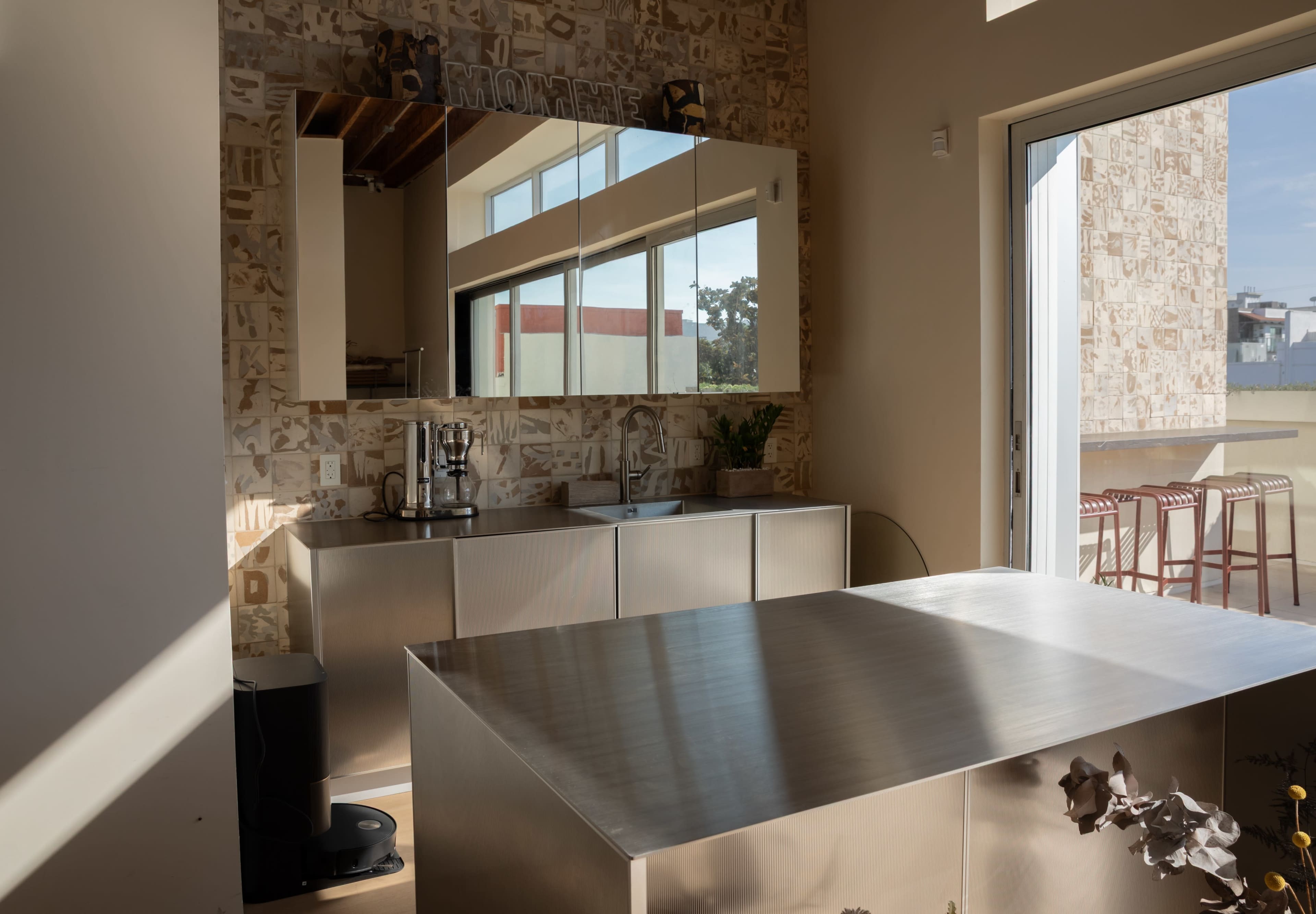 The image shows a modern kitchen with a stainless steel island, mirrored cabinets, and a large sliding glass door opening to an outdoor seating area.