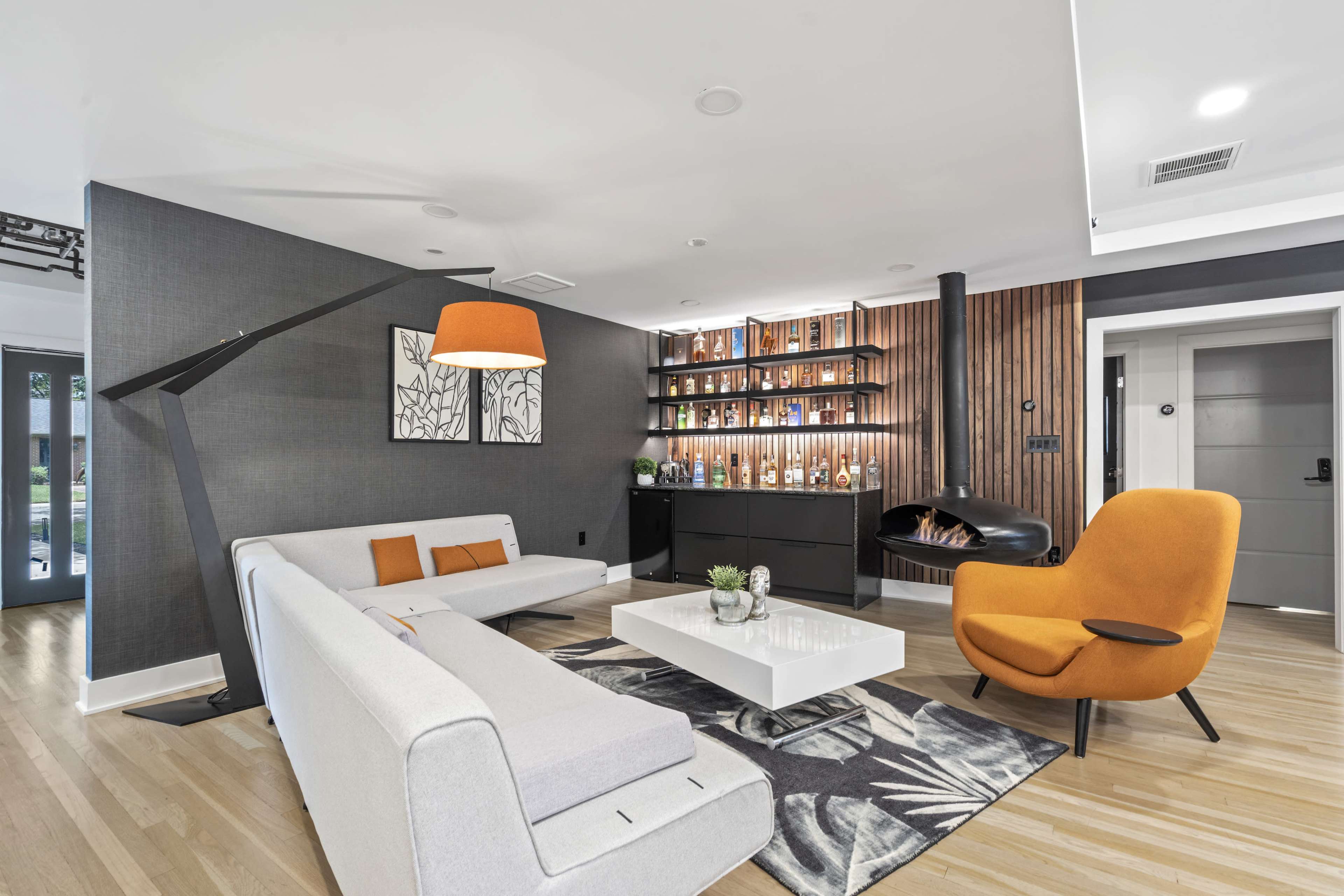 A modern living room features a gray wall, a white sofa, an orange chair, a coffee table, and a display of decorative bottles on open shelves.