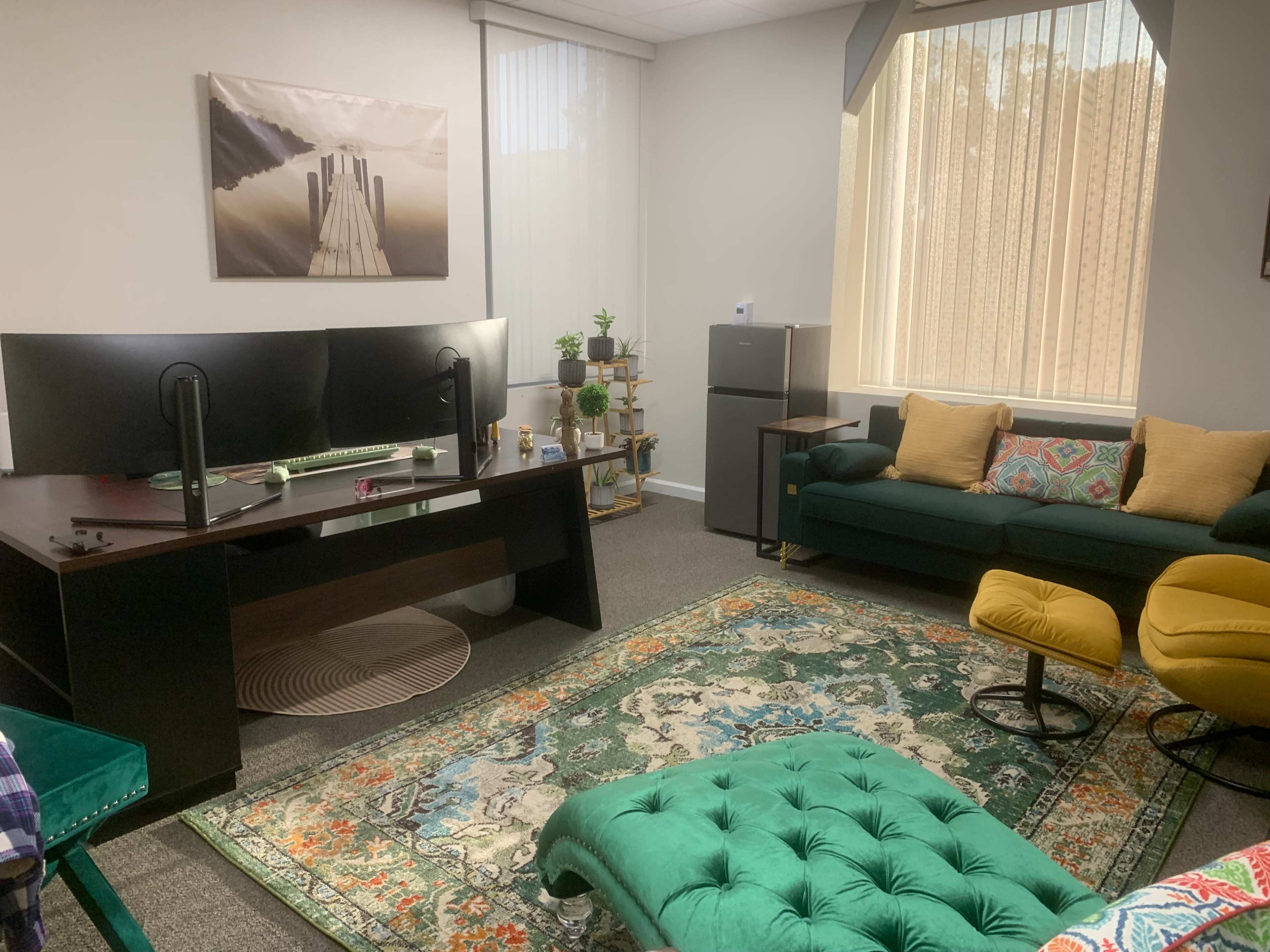 The image shows a modern office space featuring a large desk with dual monitors, a green sofa, yellow chairs, and a decorative area rug.