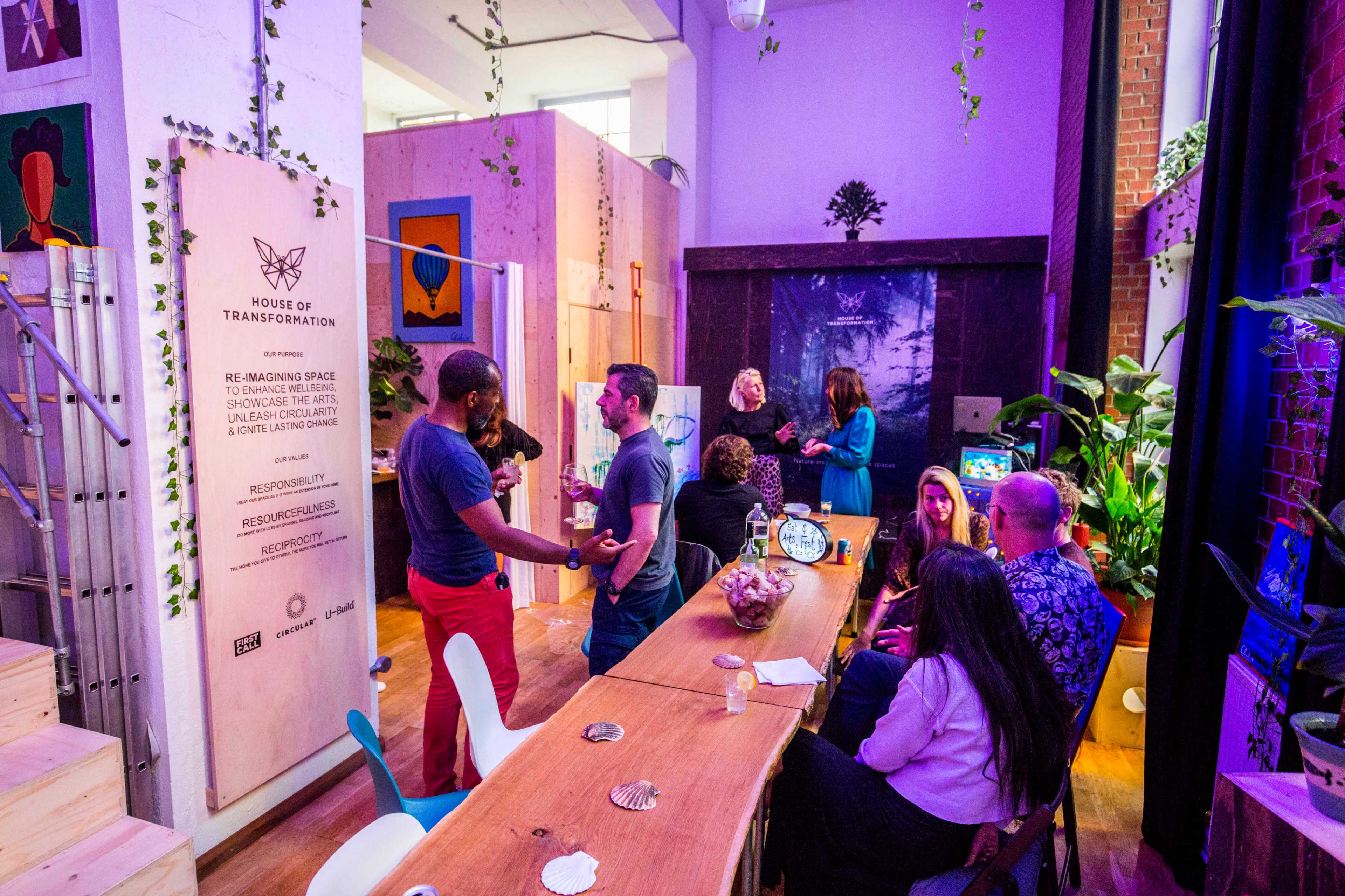 A diverse group of people engages in conversation around a long wooden table in a brightly lit, modern communal space adorned with plants and artwork on the walls.