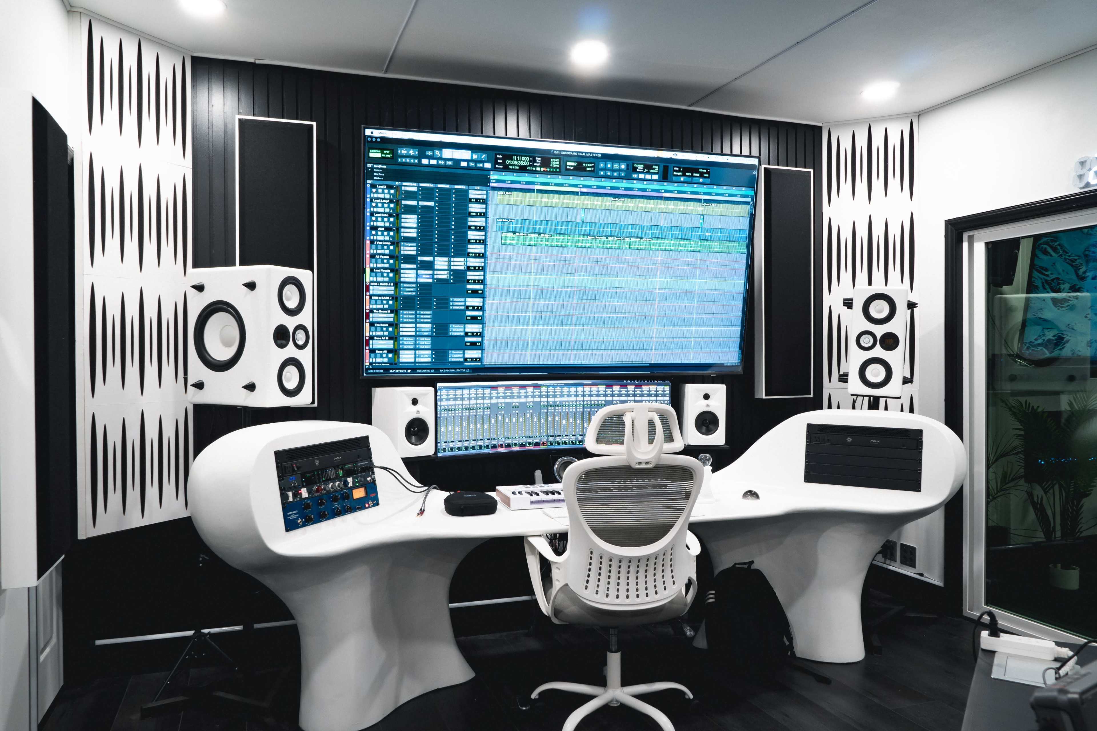 A modern music production studio features a large wall-mounted screen displaying audio tracks, flanked by speakers, with a sleek white desk and ergonomic chair in front.