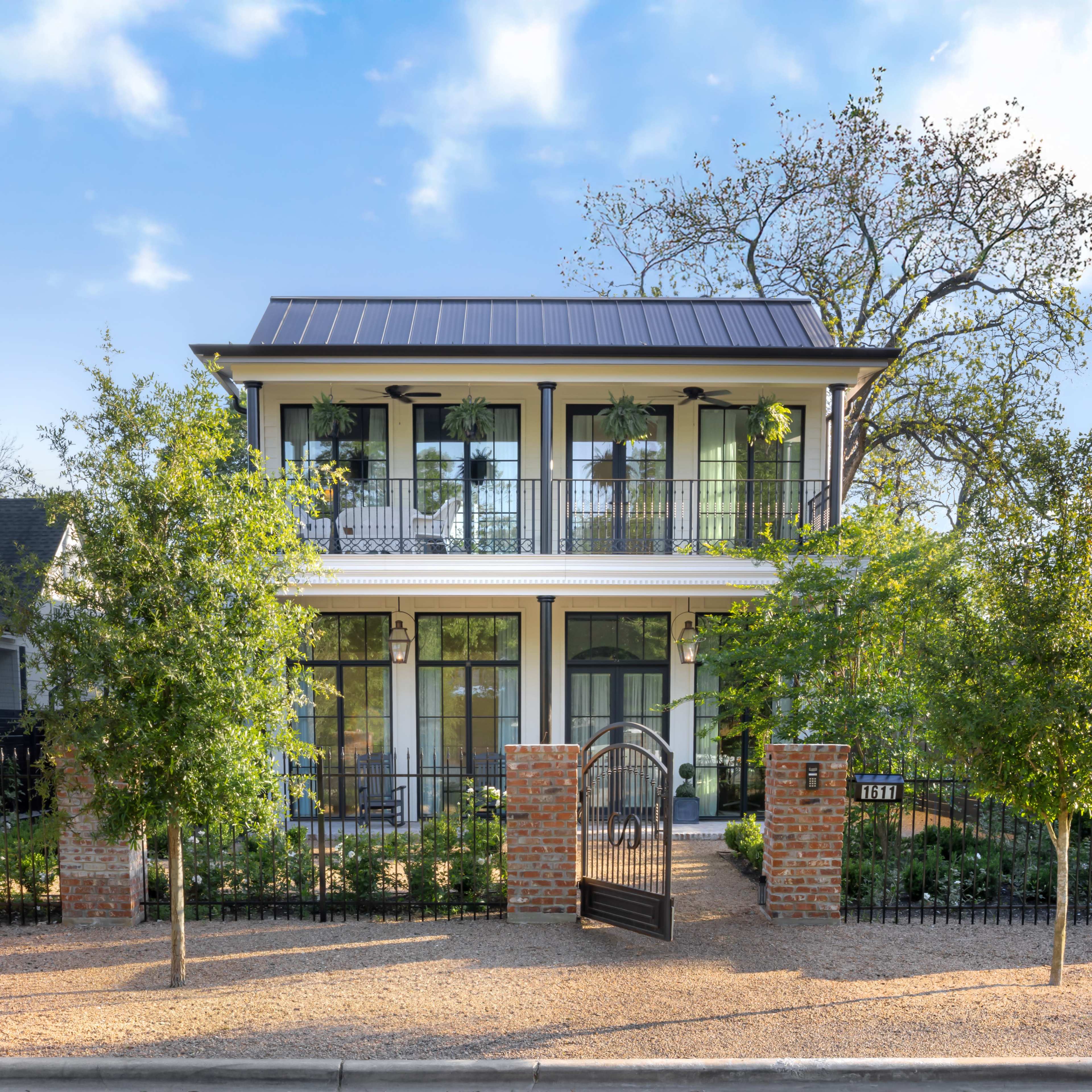 A two-story house with a black metal roof, large windows, and a front garden enclosed by a brick wall and wrought-iron gate.