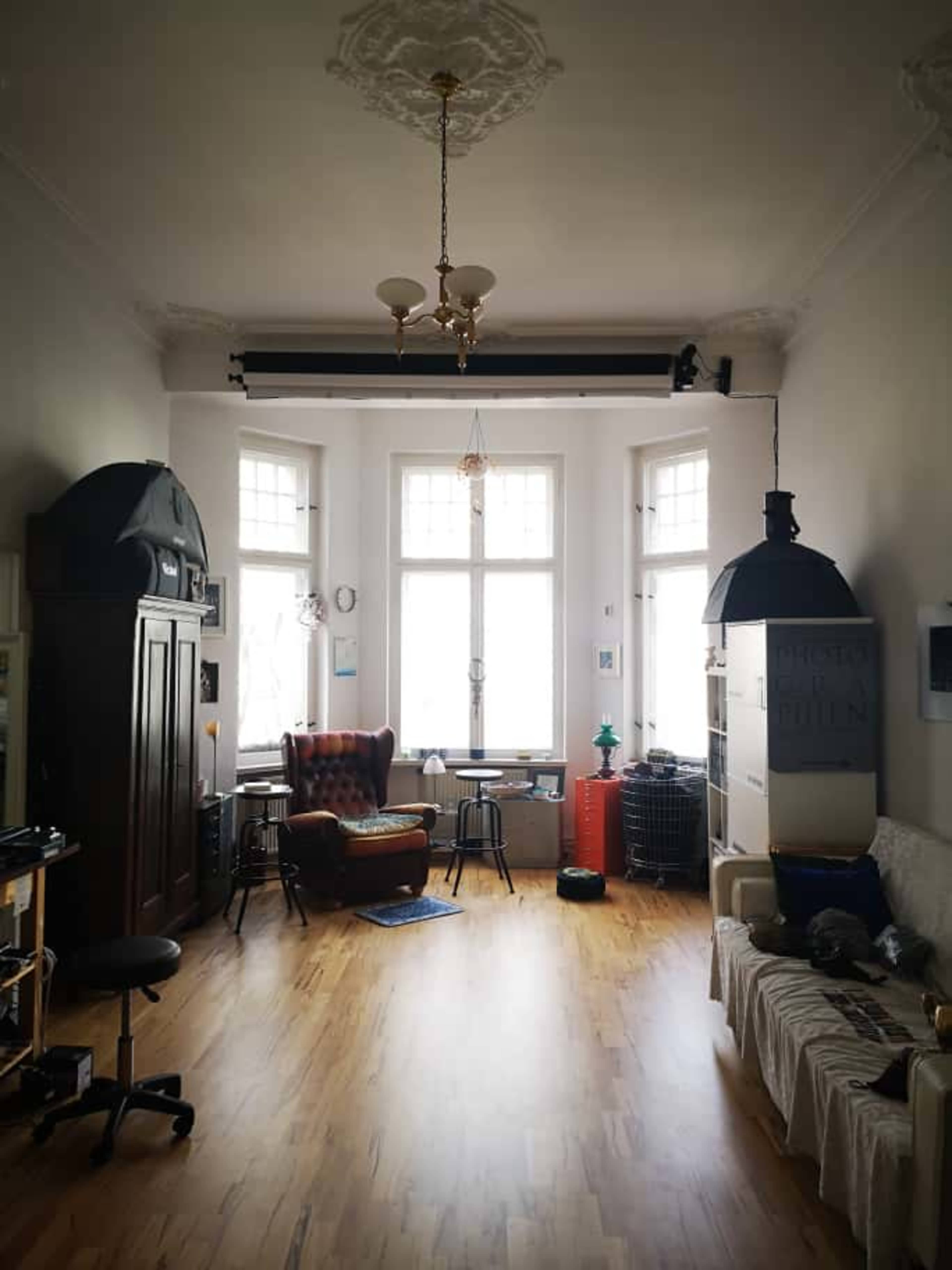 The image shows a spacious room featuring large windows, wooden flooring, a comfortable armchair, and various furniture pieces, including a cabinet and a couch.