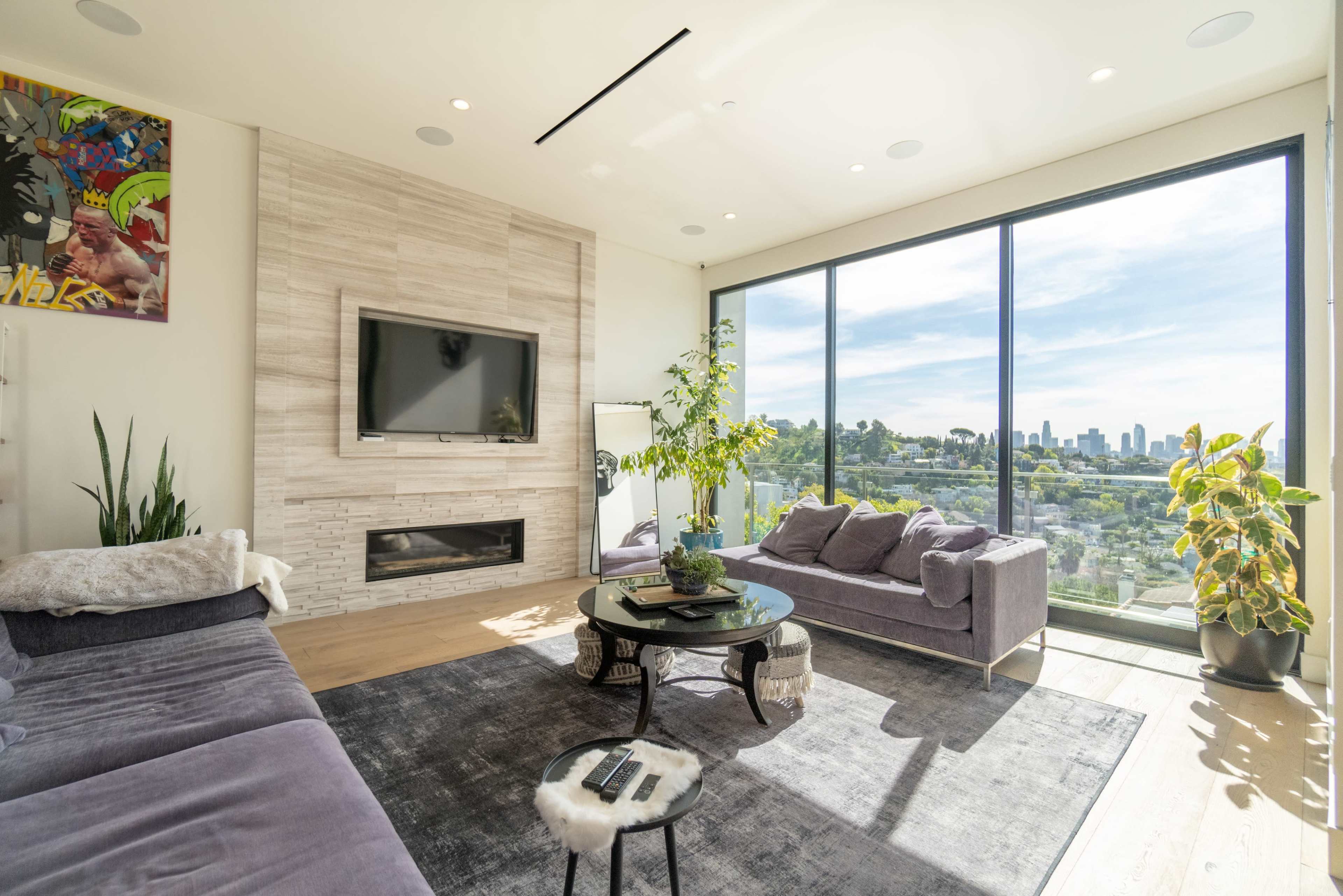A modern living room features a large window with a city view, a wall-mounted TV, a fireplace, and seating arranged around a circular coffee table.