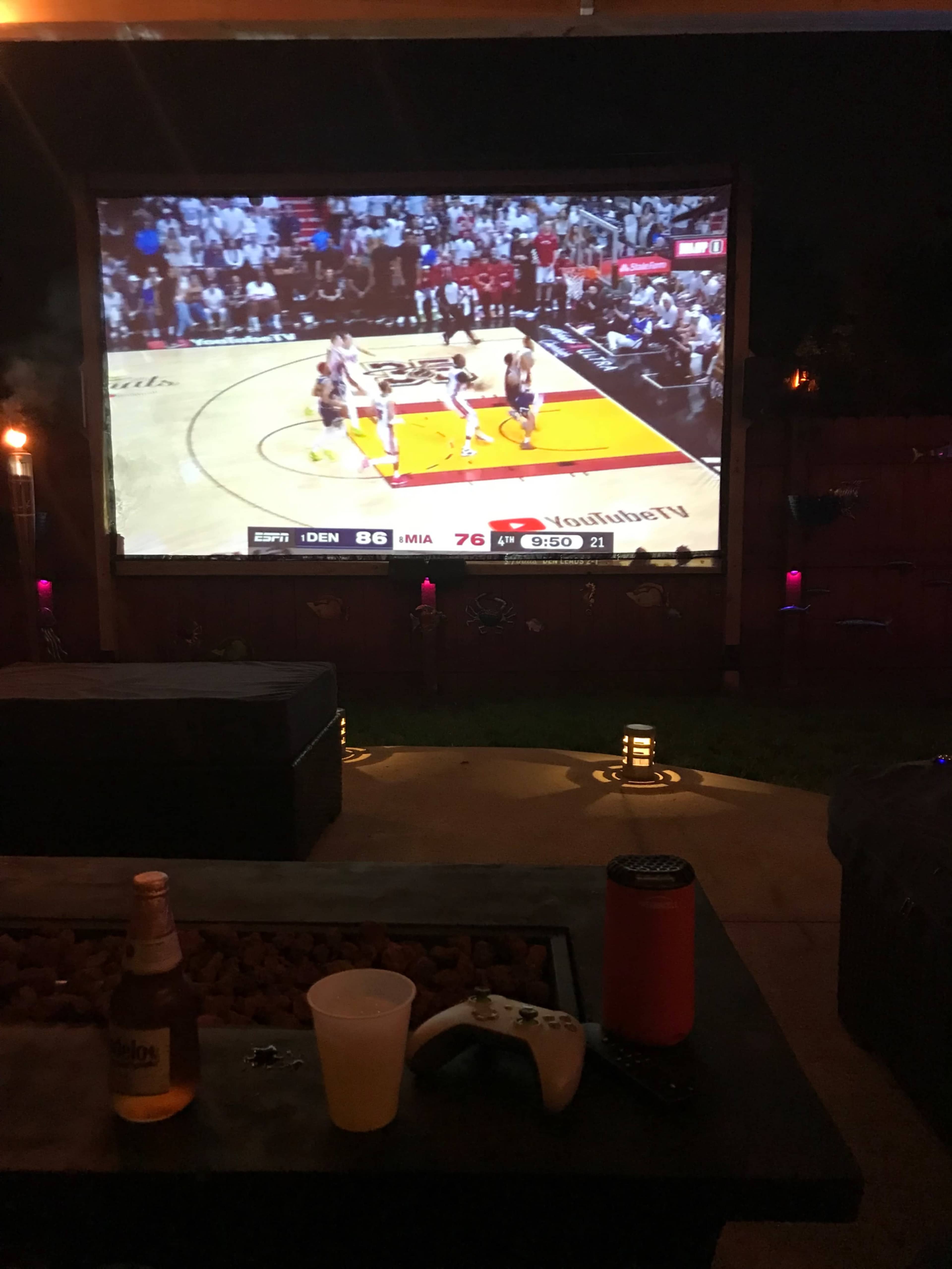 A large outdoor screen displays a basketball game, with a beverage and gaming console nearby in a well-lit backyard setting.
