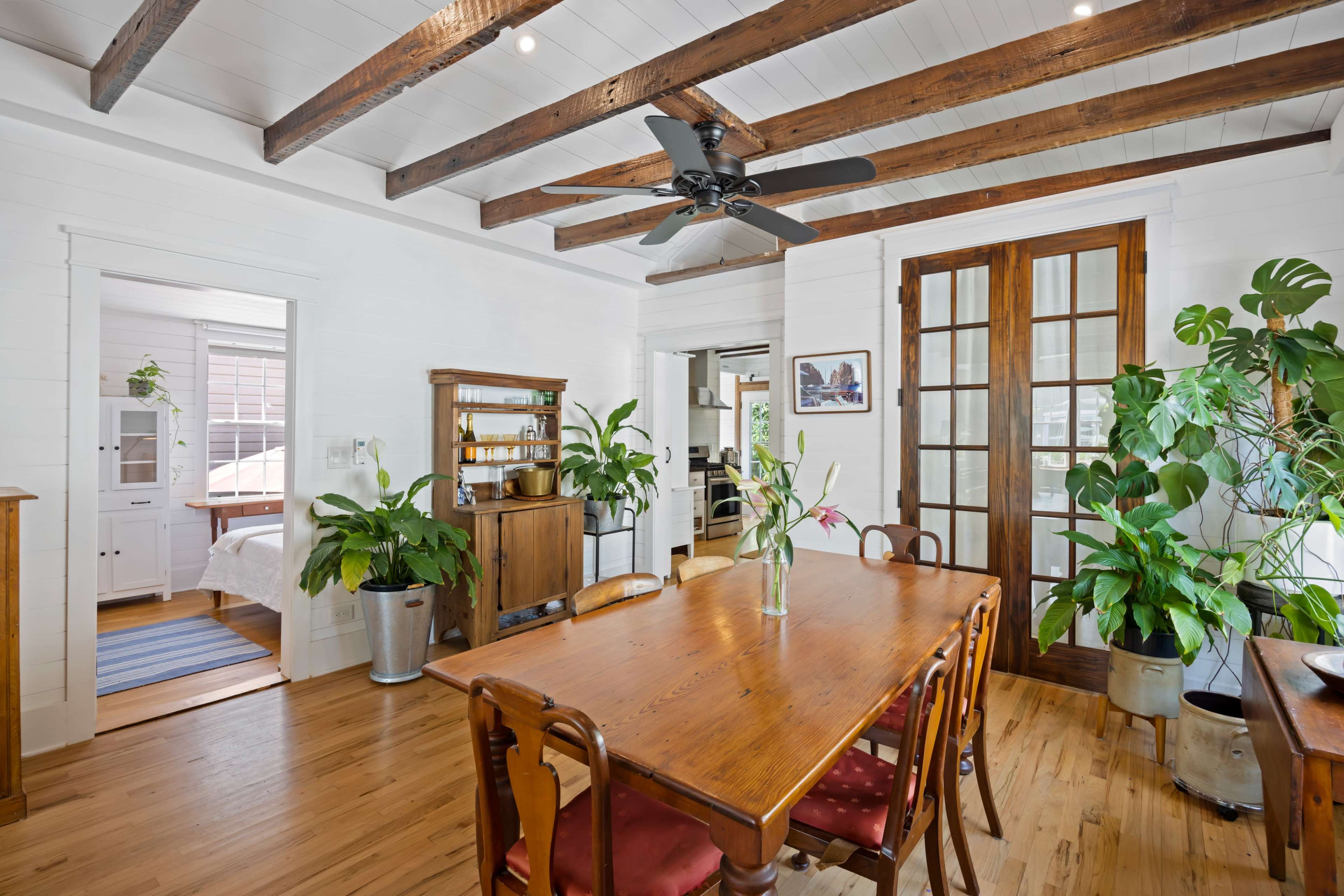 A dining area features a wooden table surrounded by chairs, with potted plants and wooden furniture, under exposed beams and a ceiling fan.
