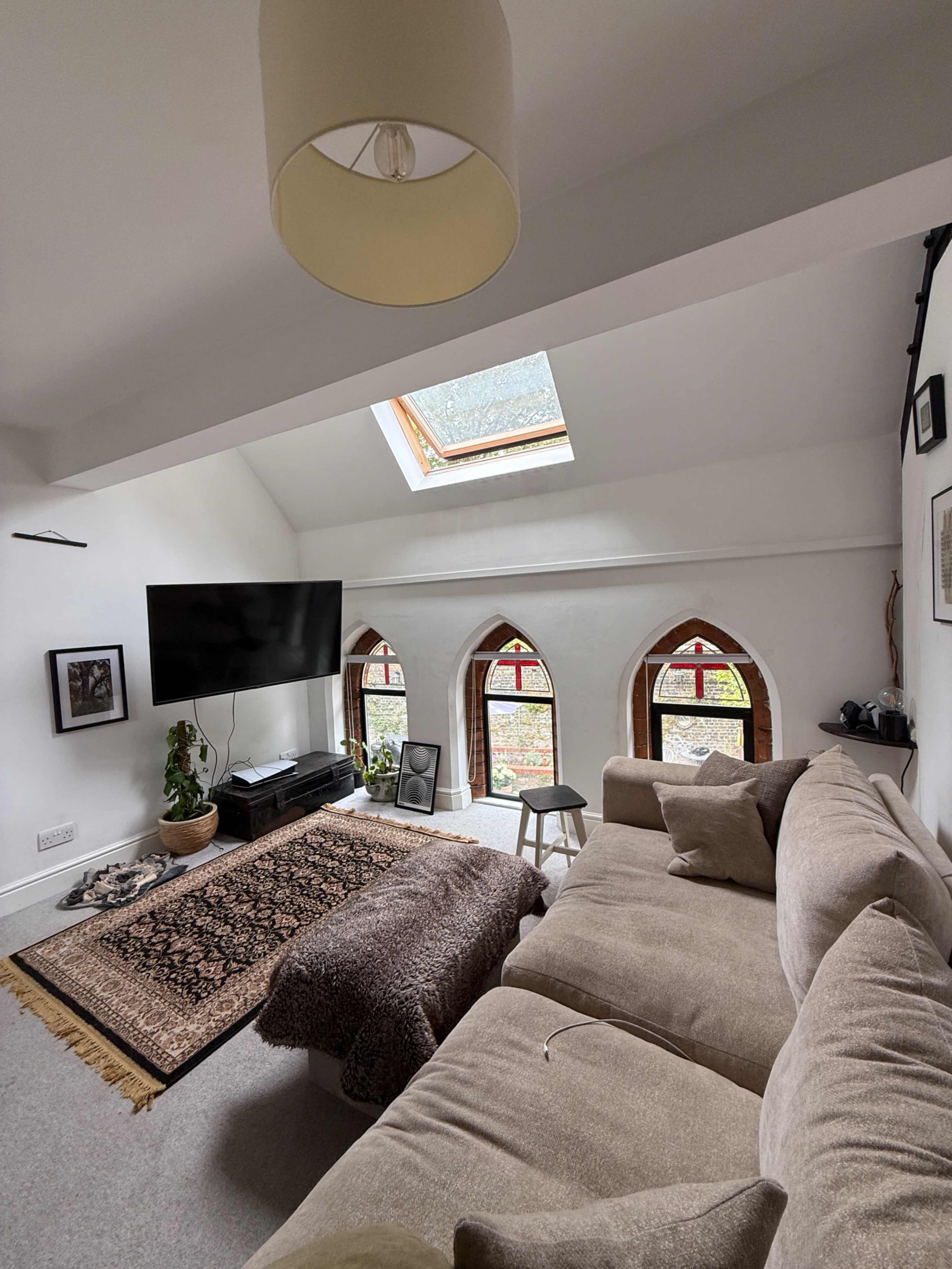A cozy living room features a sectional sofa, a television, and three arched windows with colored glass, illuminated by natural light from a skylight.
