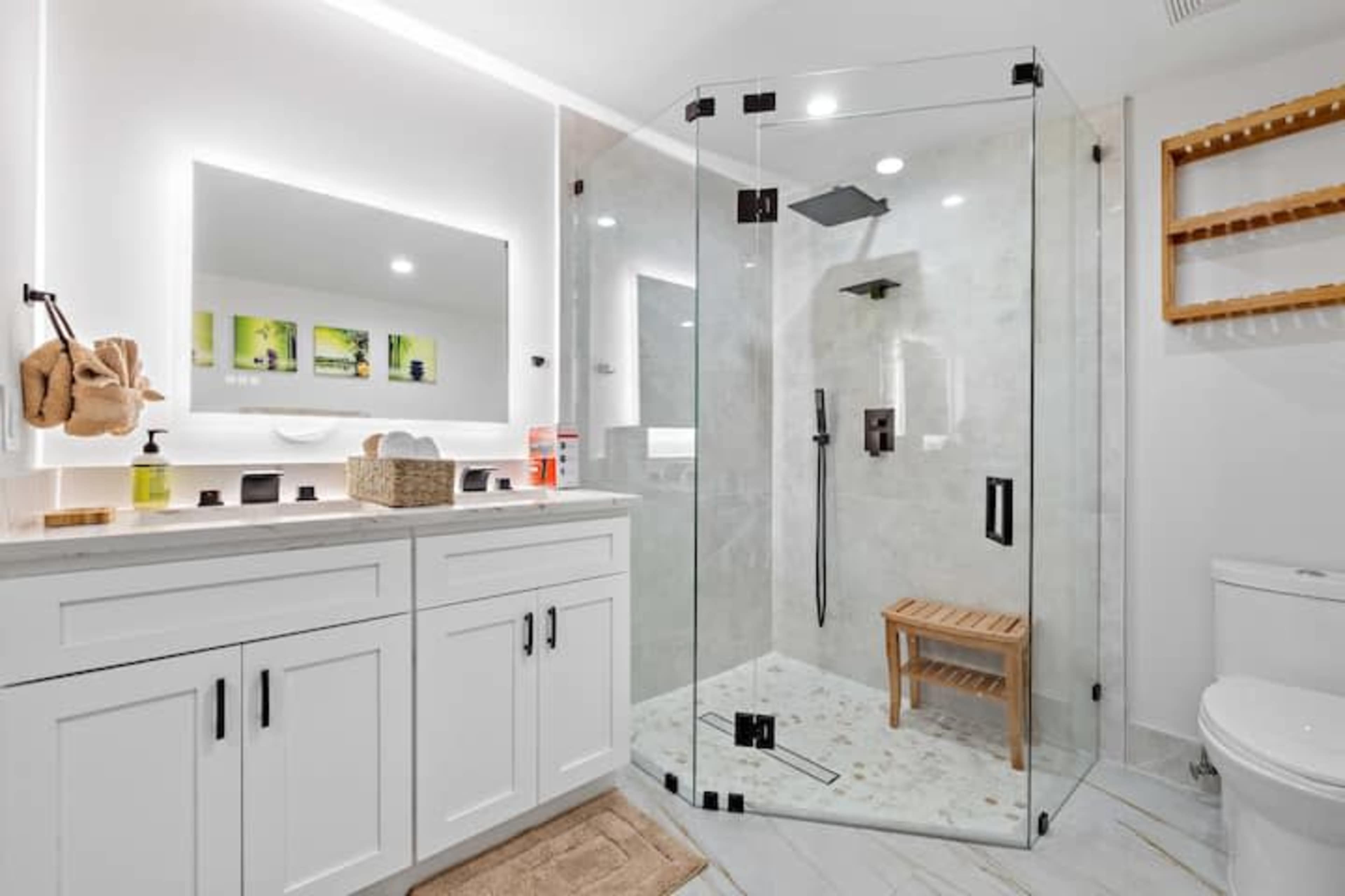 The image shows a modern bathroom featuring a glass-enclosed shower, a double vanity with black hardware, and a wooden shower stool.