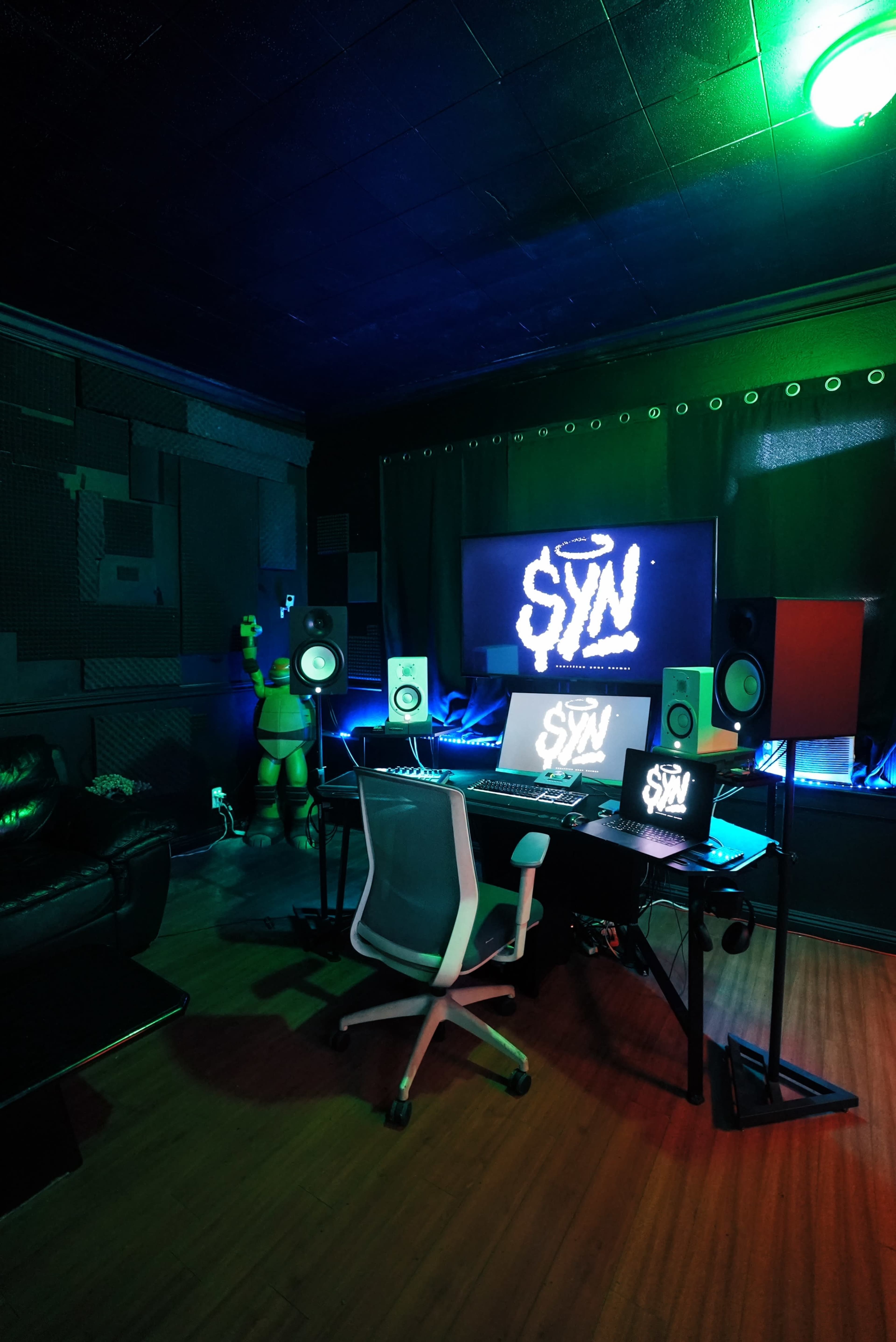 The image shows a modern music studio with a desk, computer, speakers, and colorful lighting.