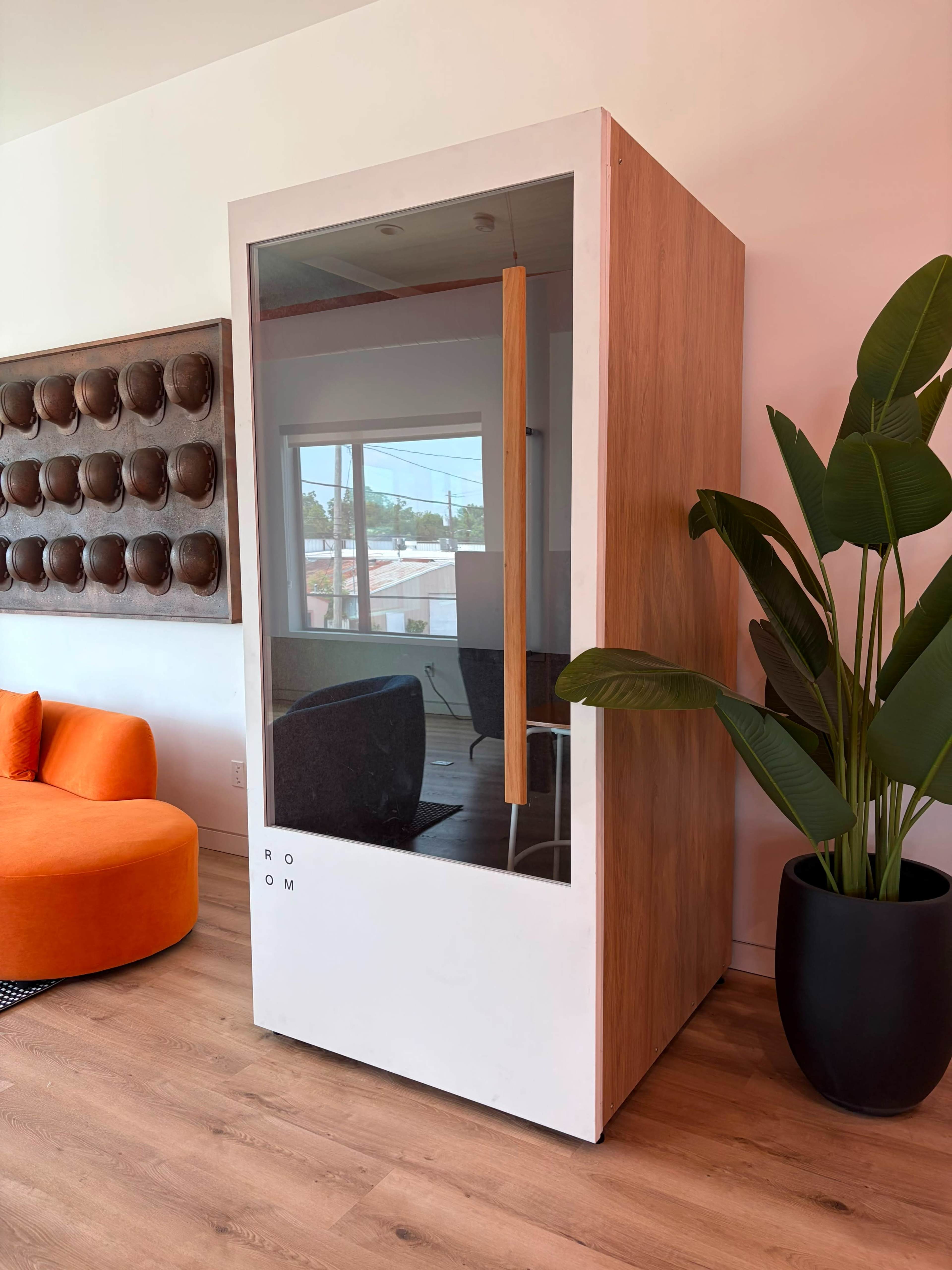 The image shows a modern phone booth with a wooden frame and glass front positioned next to a large green plant and an orange sofa.
