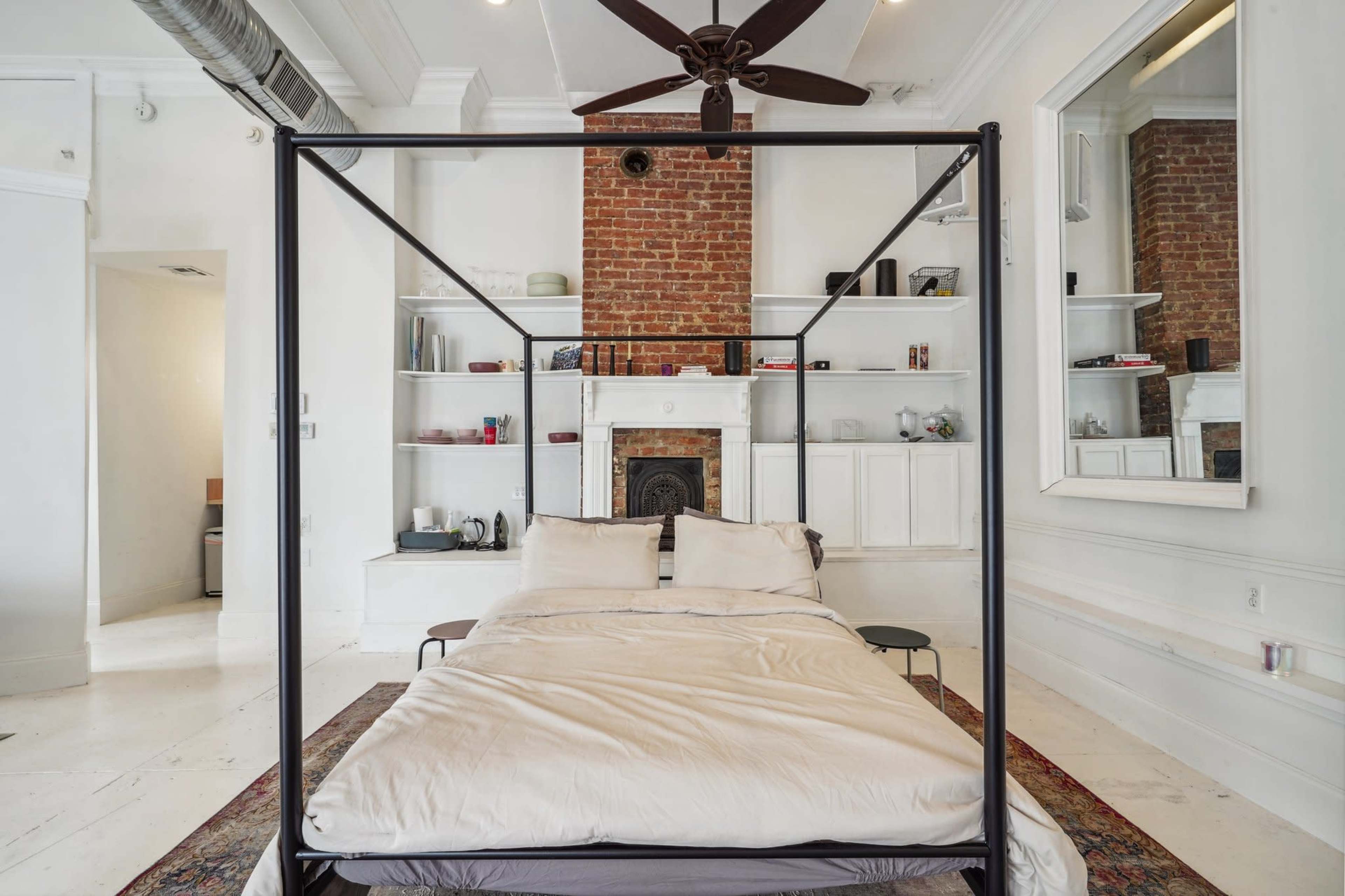A four-poster bed is positioned in a spacious room featuring exposed brick walls, a fireplace, and built-in shelves filled with books and decor.