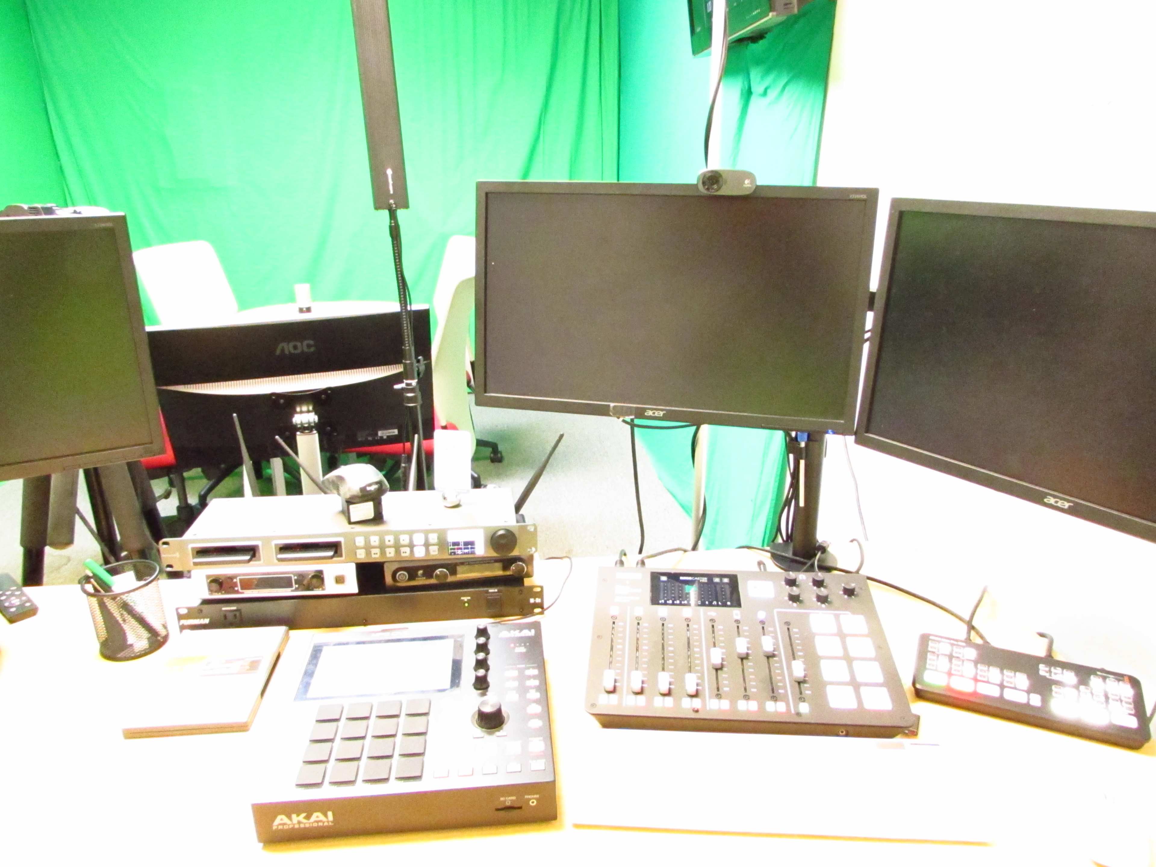 The image shows a production setup featuring two monitors, a mixer, a drum pad, and additional equipment, set against a green backdrop.