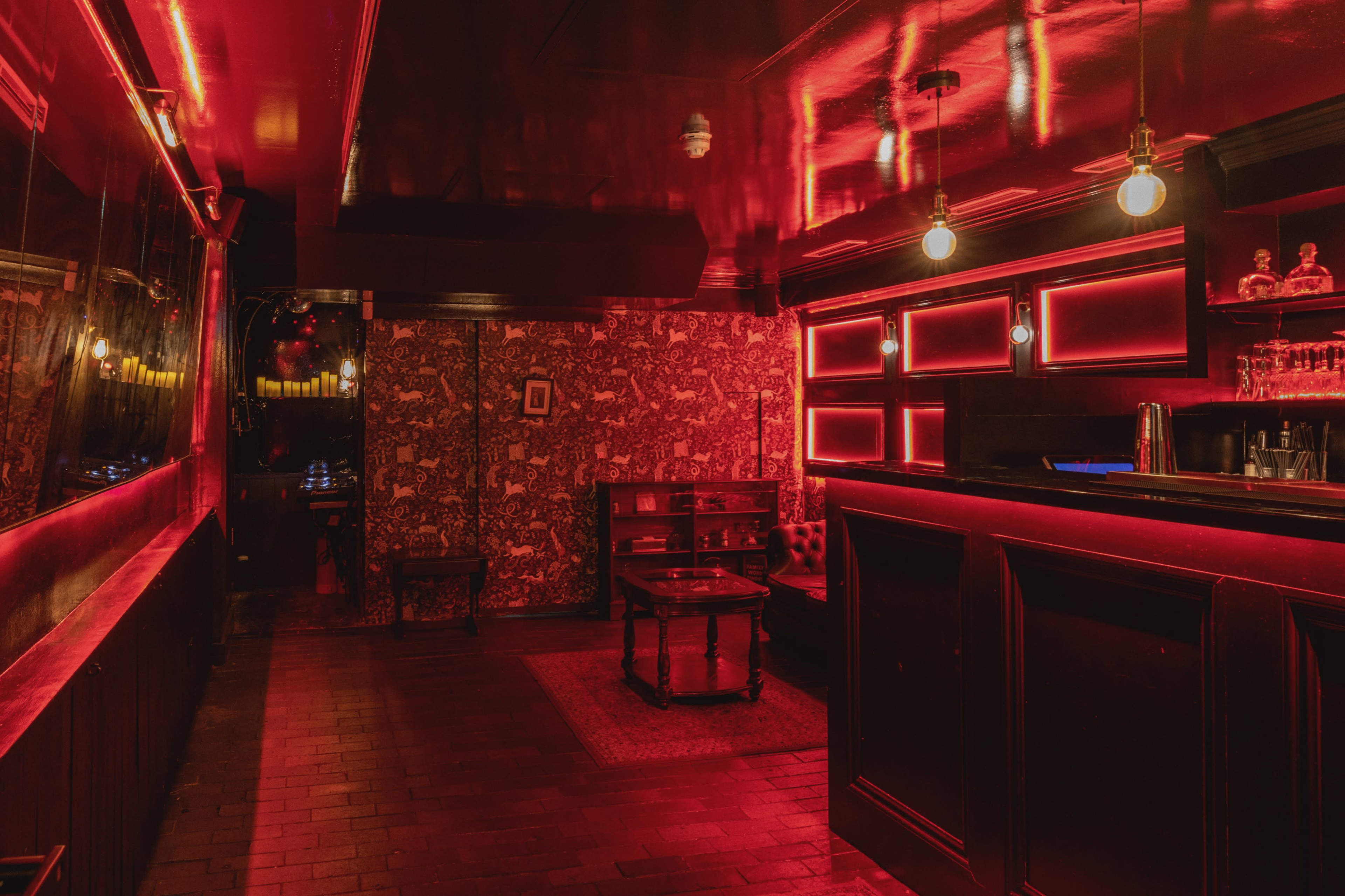 A dimly lit bar interior with red lighting, wooden flooring, and decorative wallpaper.