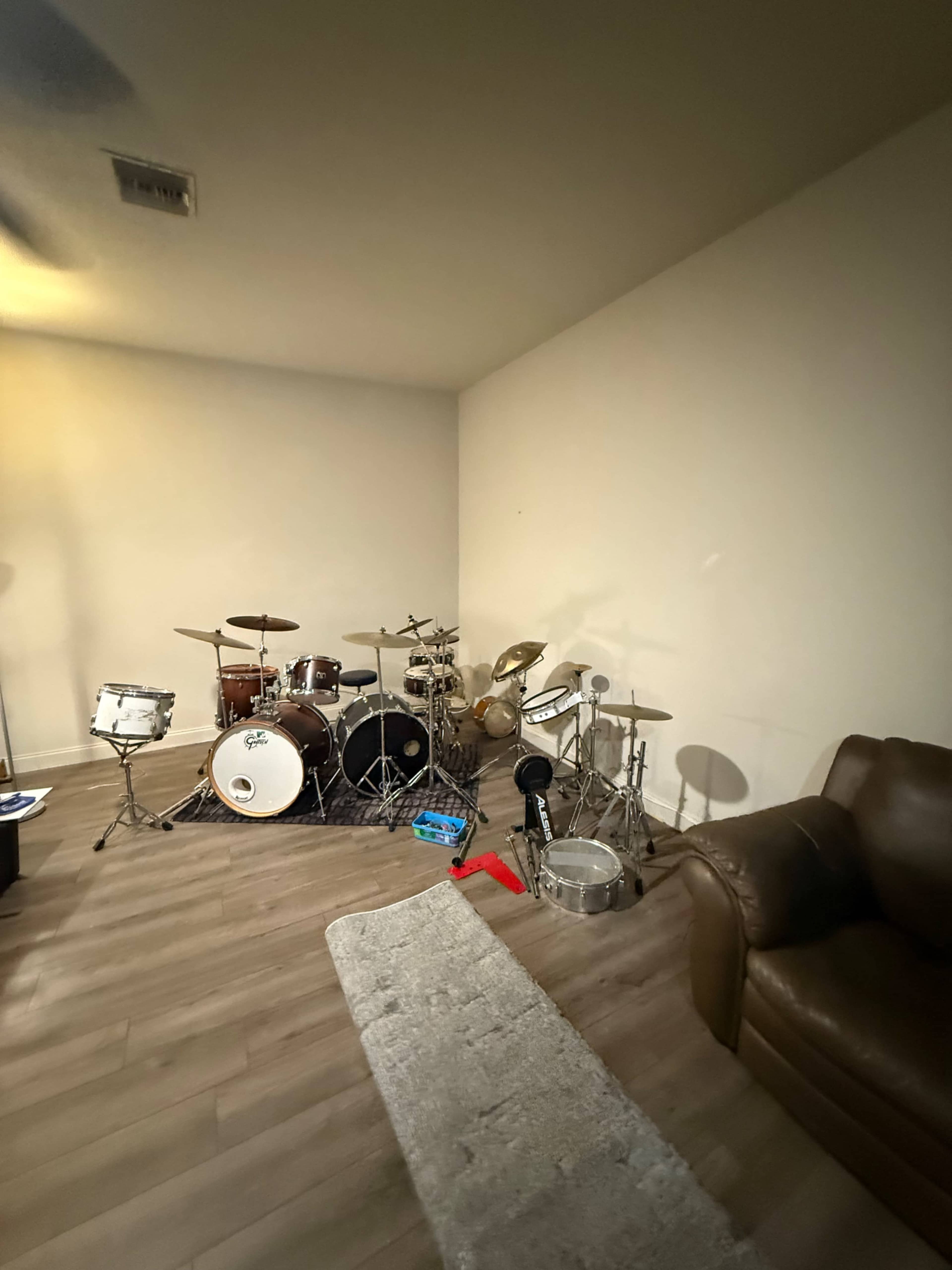 The image shows a small room featuring a drum set with various drums and cymbals, a couch, and a small rug on the floor.