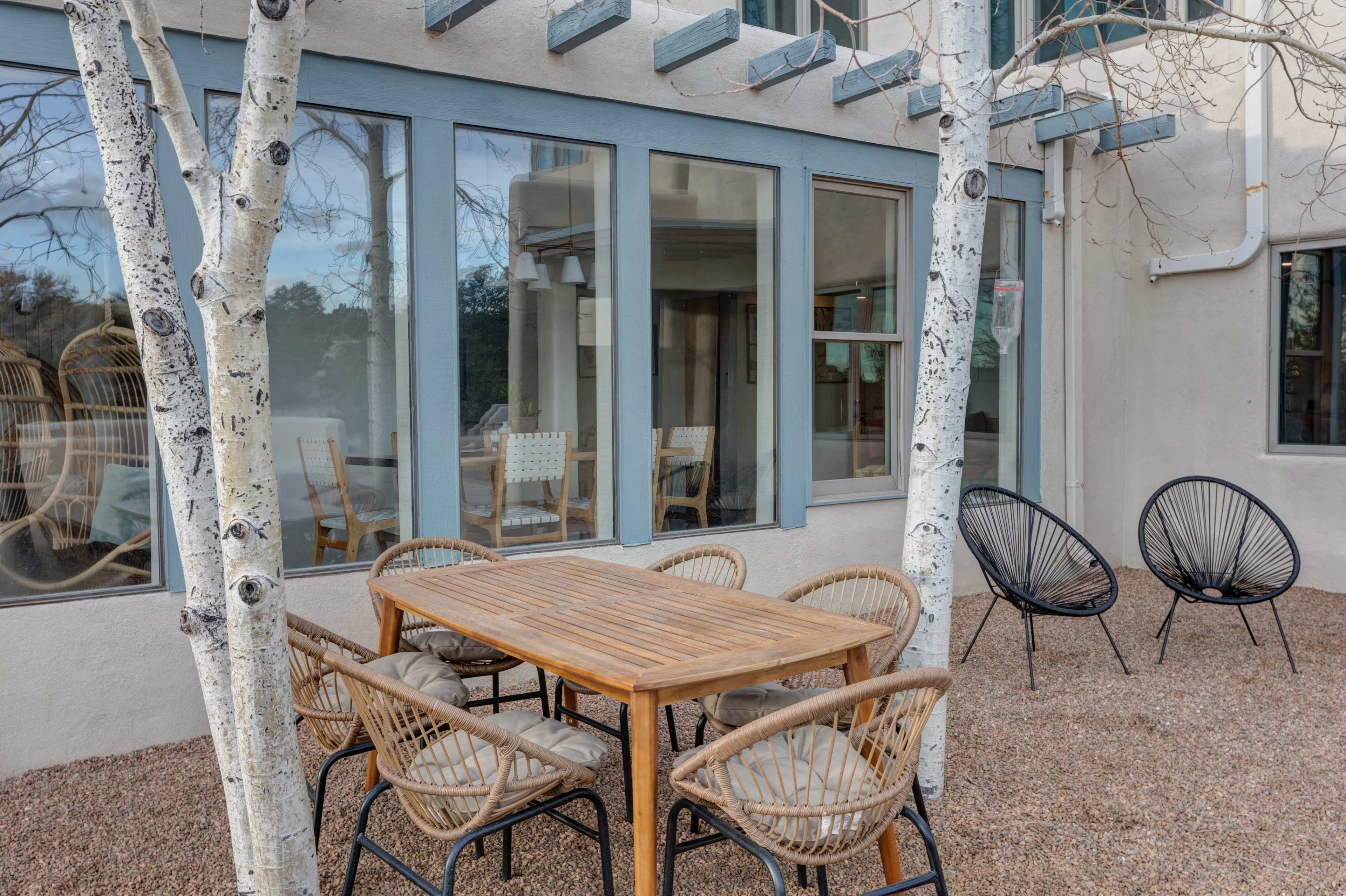A wooden table with six rattan chairs is positioned on a gravel patio beside two white birch trees, adjacent to a building featuring large windows.