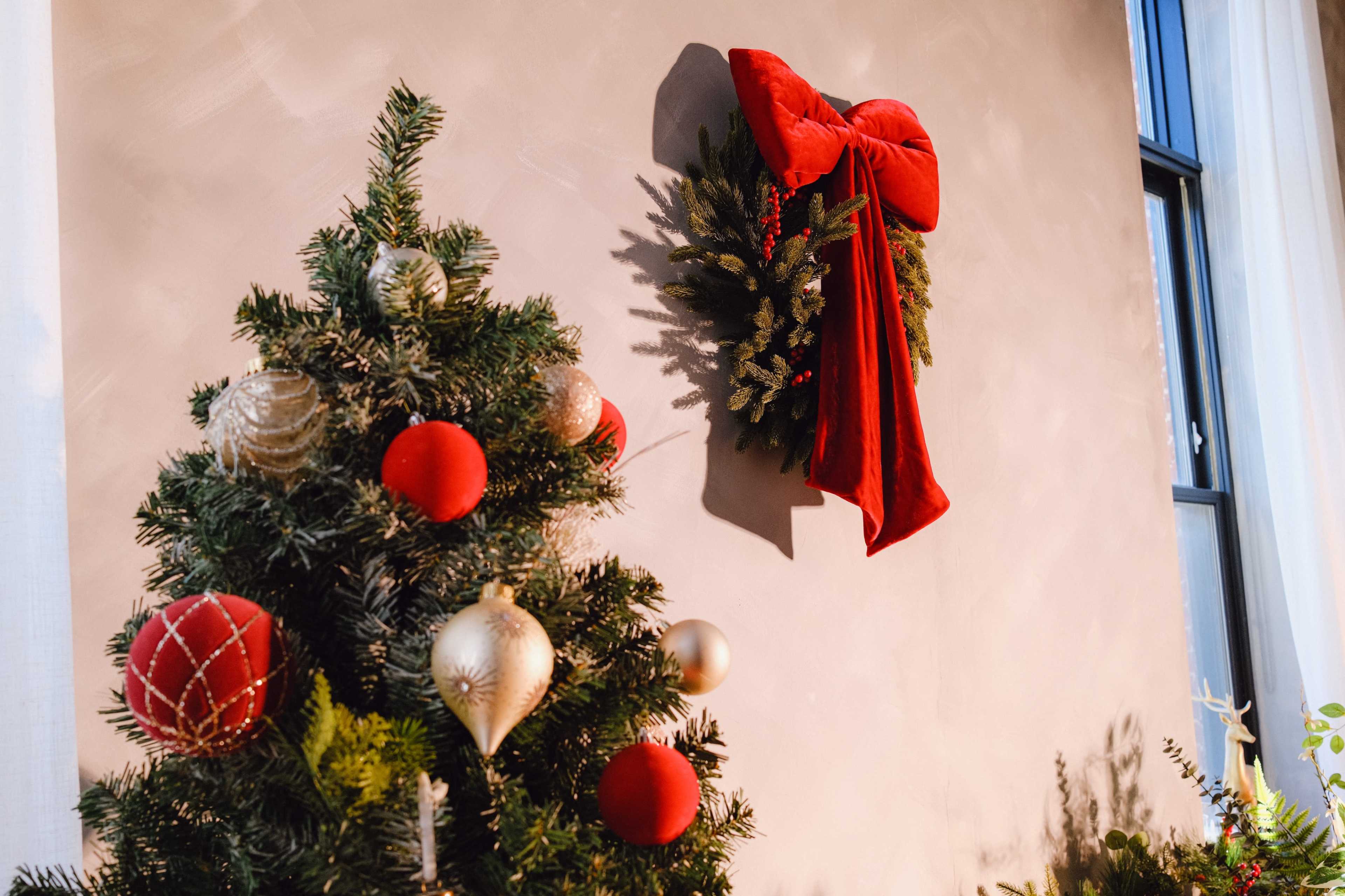 A decorated Christmas tree with red and gold ornaments is positioned next to a wall featuring a red bow-adorned wreath.