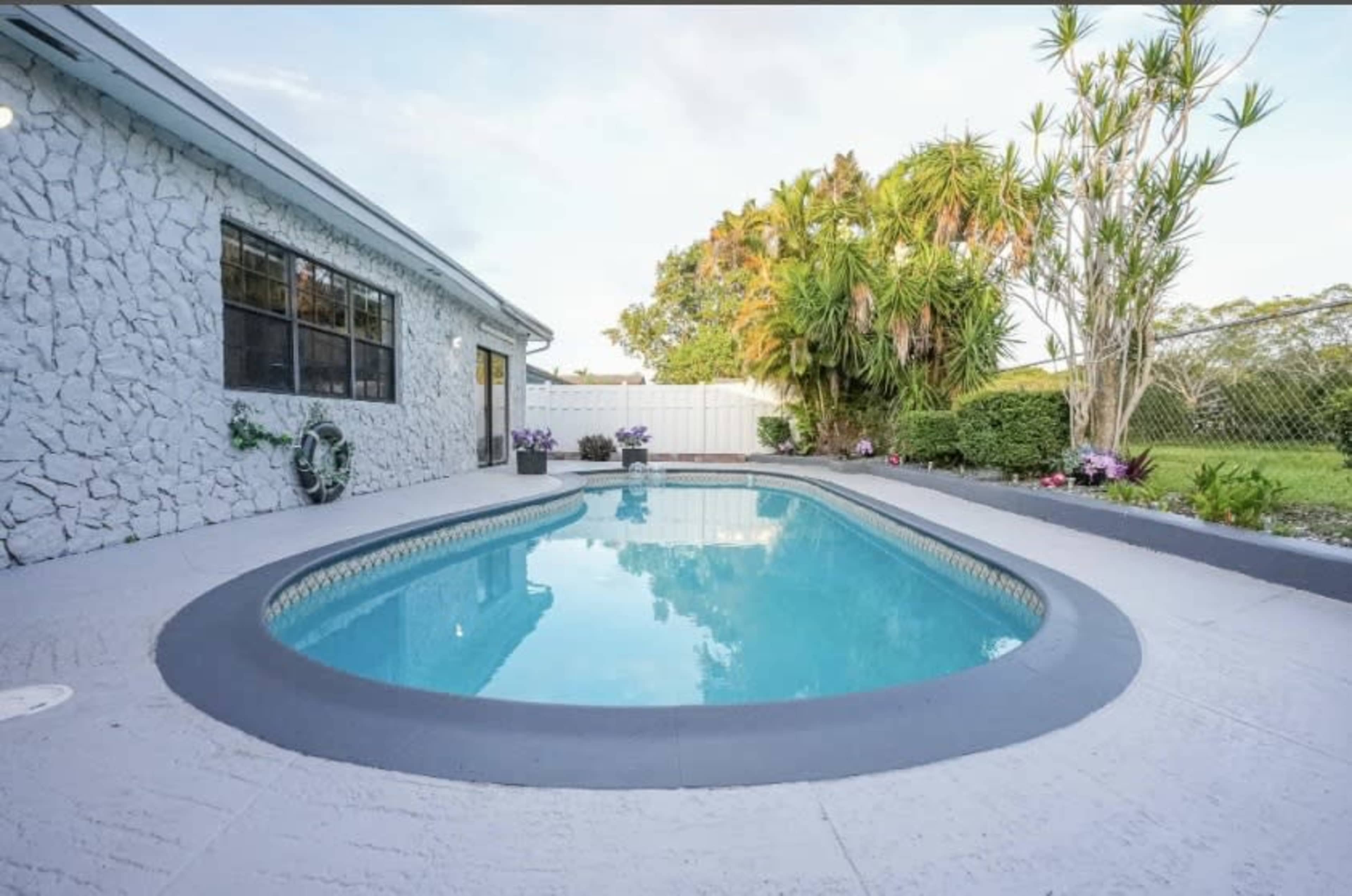 The image shows a swimming pool surrounded by a landscaped yard with tropical plants and a white house in the background.