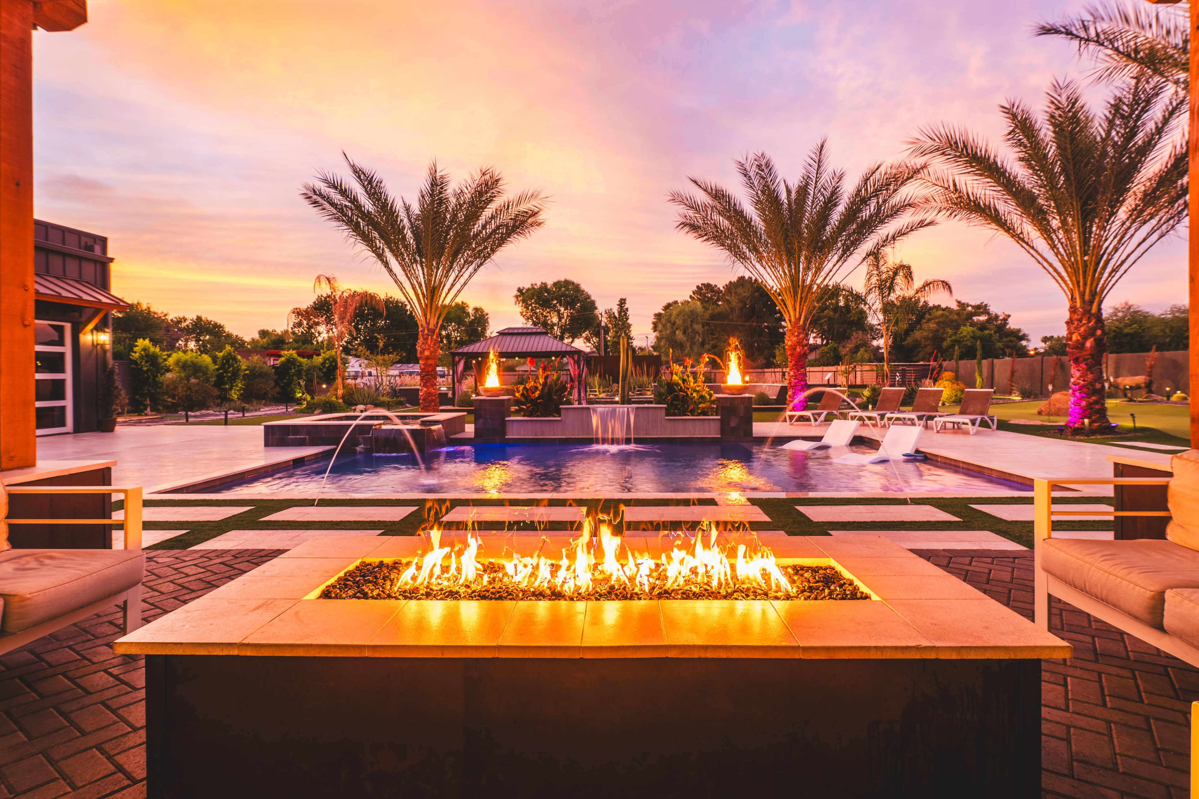 The image depicts a backyard oasis featuring a swimming pool, palm trees, and a fire pit, all set against a colorful sunset.