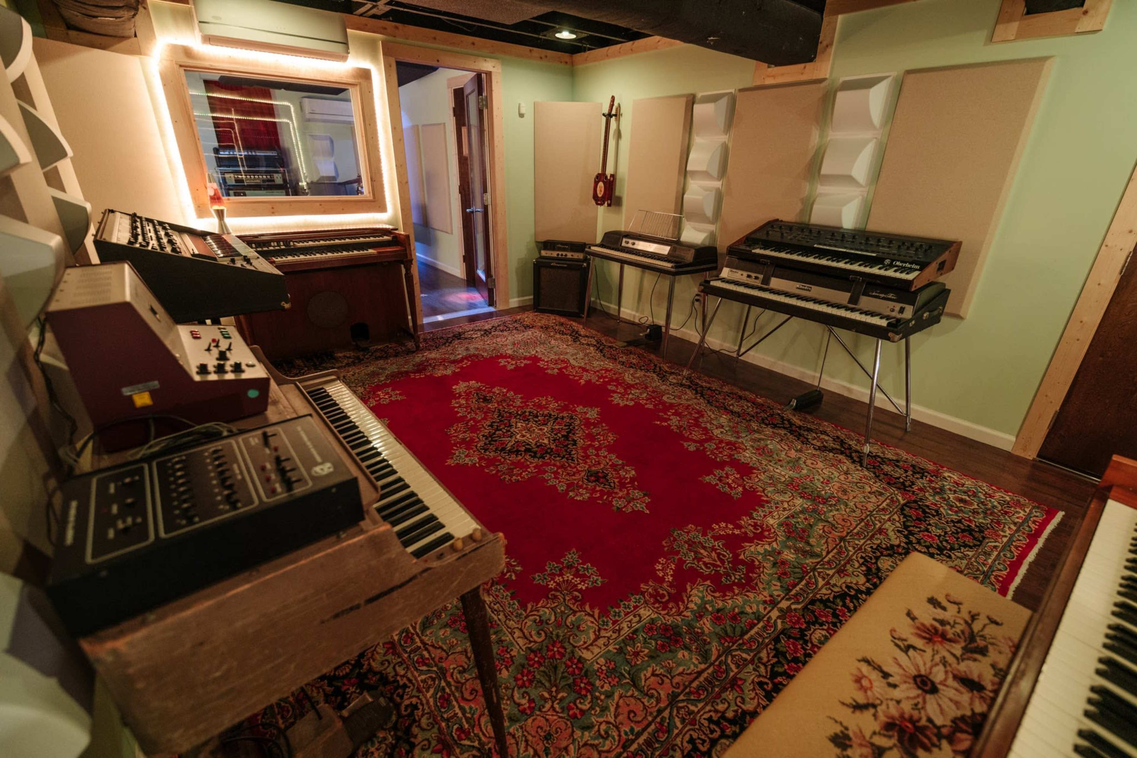 A music studio featuring various synthesizers and keyboards surrounded by plush carpeting and acoustic panels.