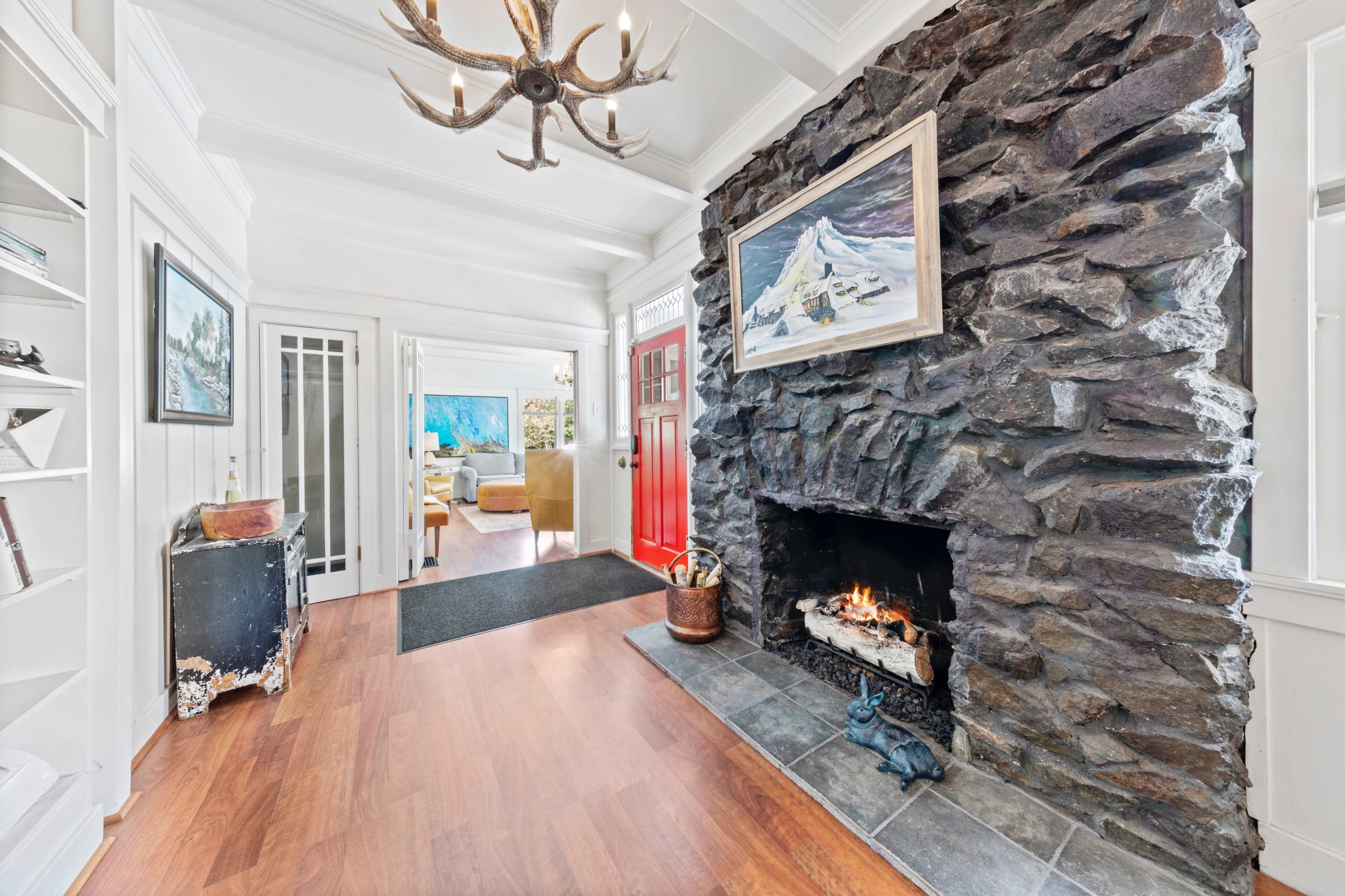 A home interior featuring a large stone fireplace, a red front door, and a view into a bright living area with artwork and furniture.