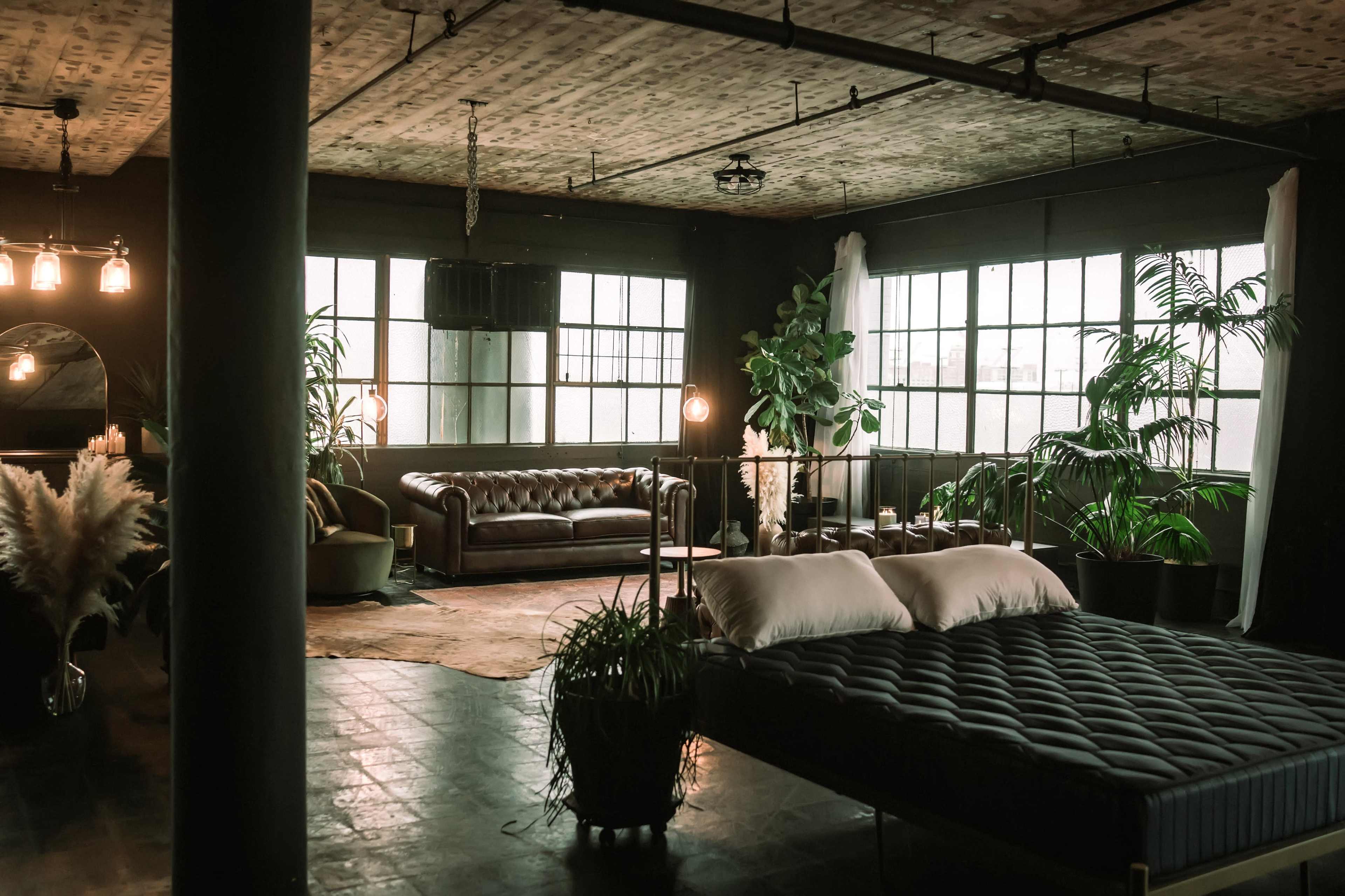 The image features a spacious, dimly lit industrial-style room with large windows, a bed with a dark mattress, a leather sofa, and various indoor plants.