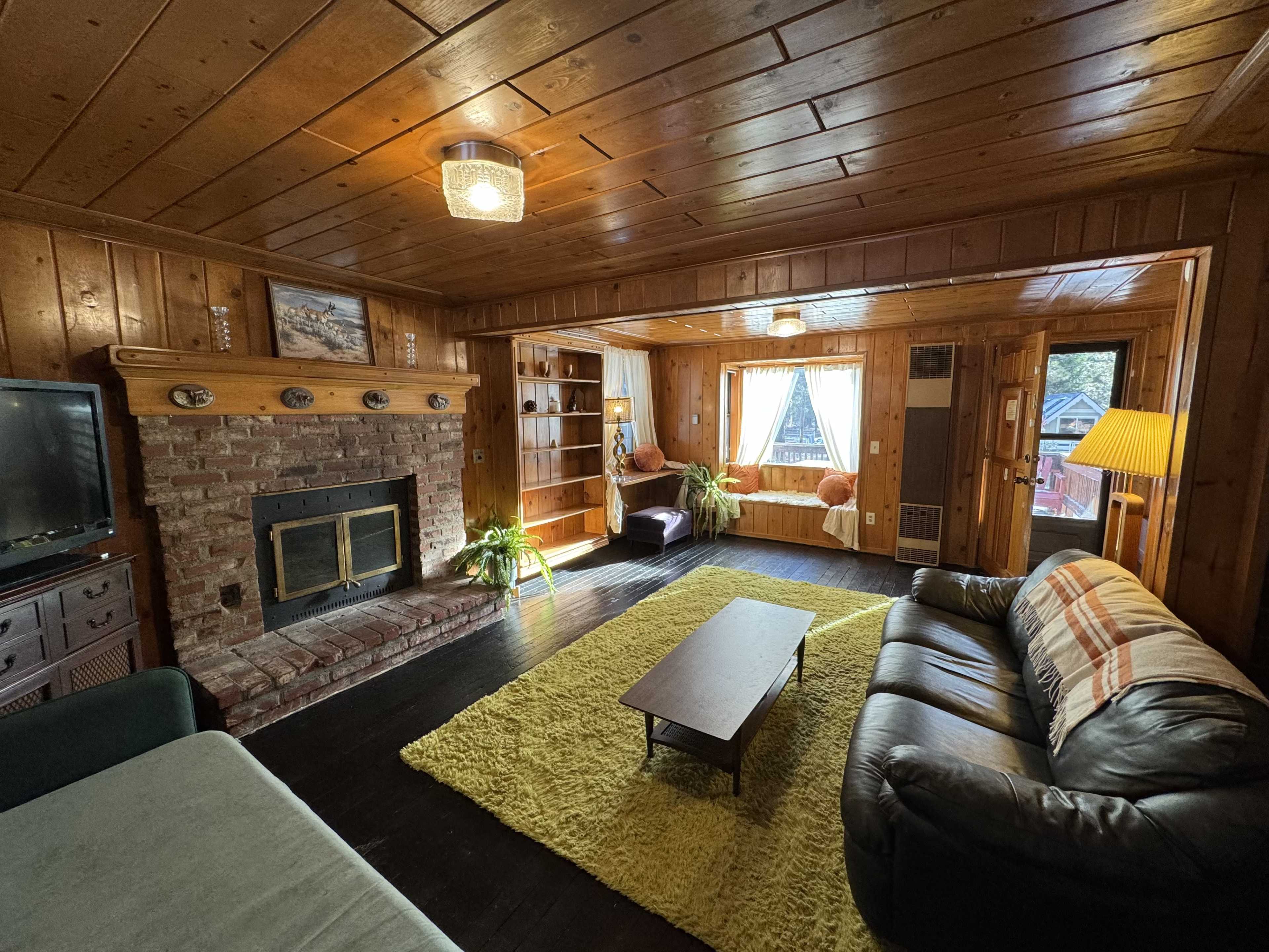 A rustic living room features a brick fireplace, wooden walls, a dark leather sofa, and a coffee table on a green rug.