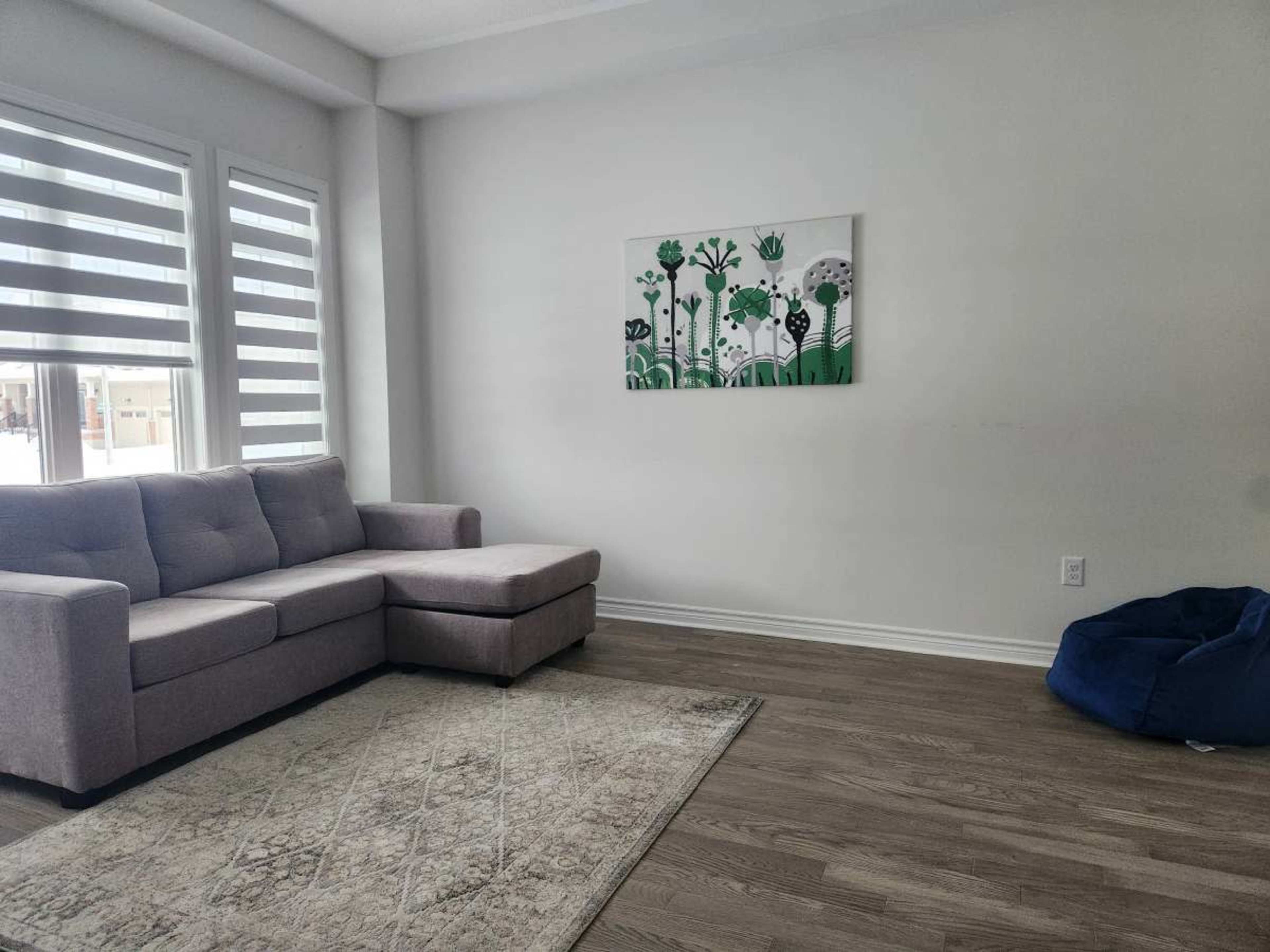 A light-filled living room features a gray sectional sofa, a wall art piece with green accents, and a blue bean bag chair on a wooden floor.