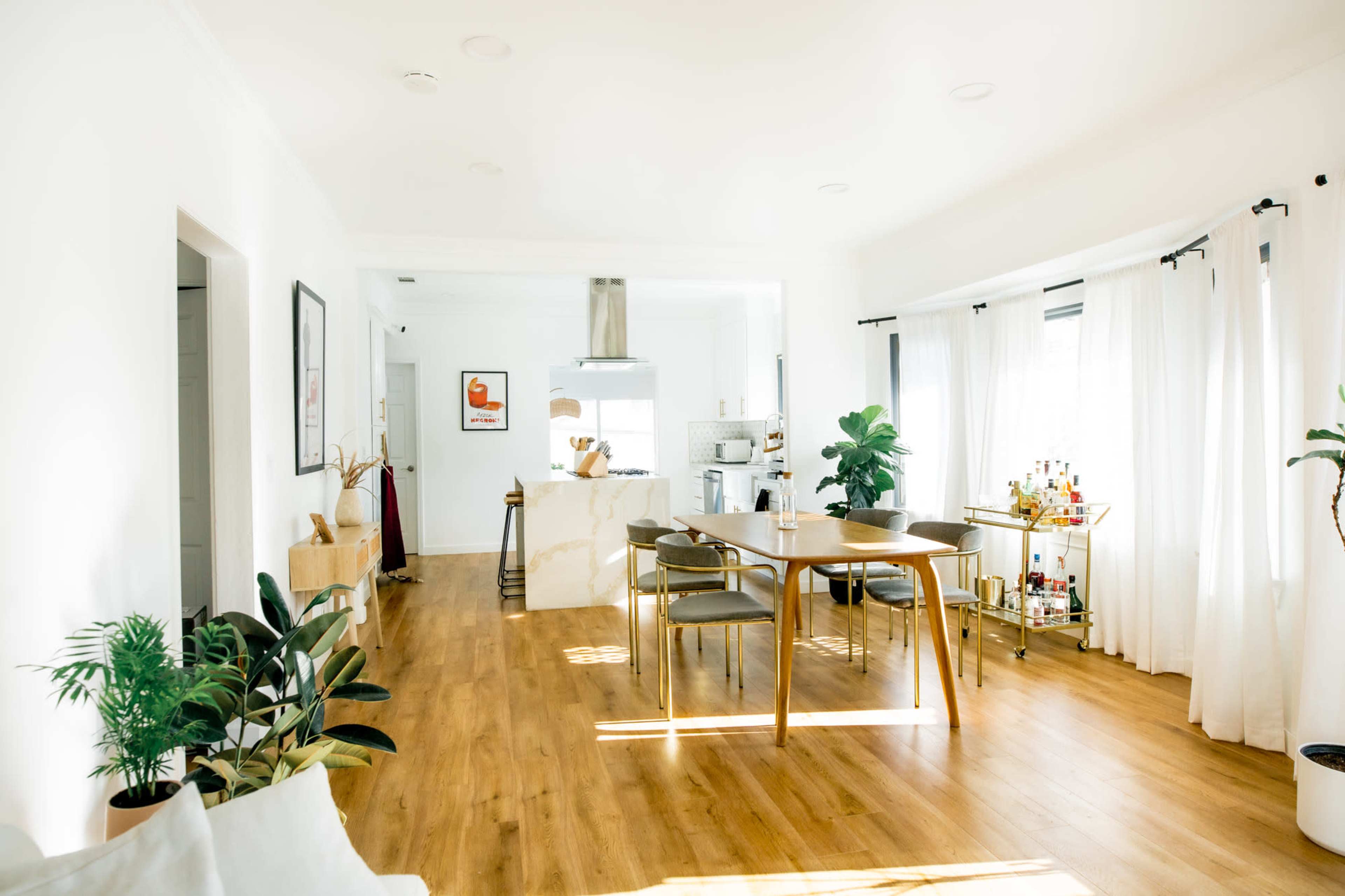 A bright, modern open-concept living space features a dining area with a wooden table, surrounded by chairs, and a kitchen visible in the background.