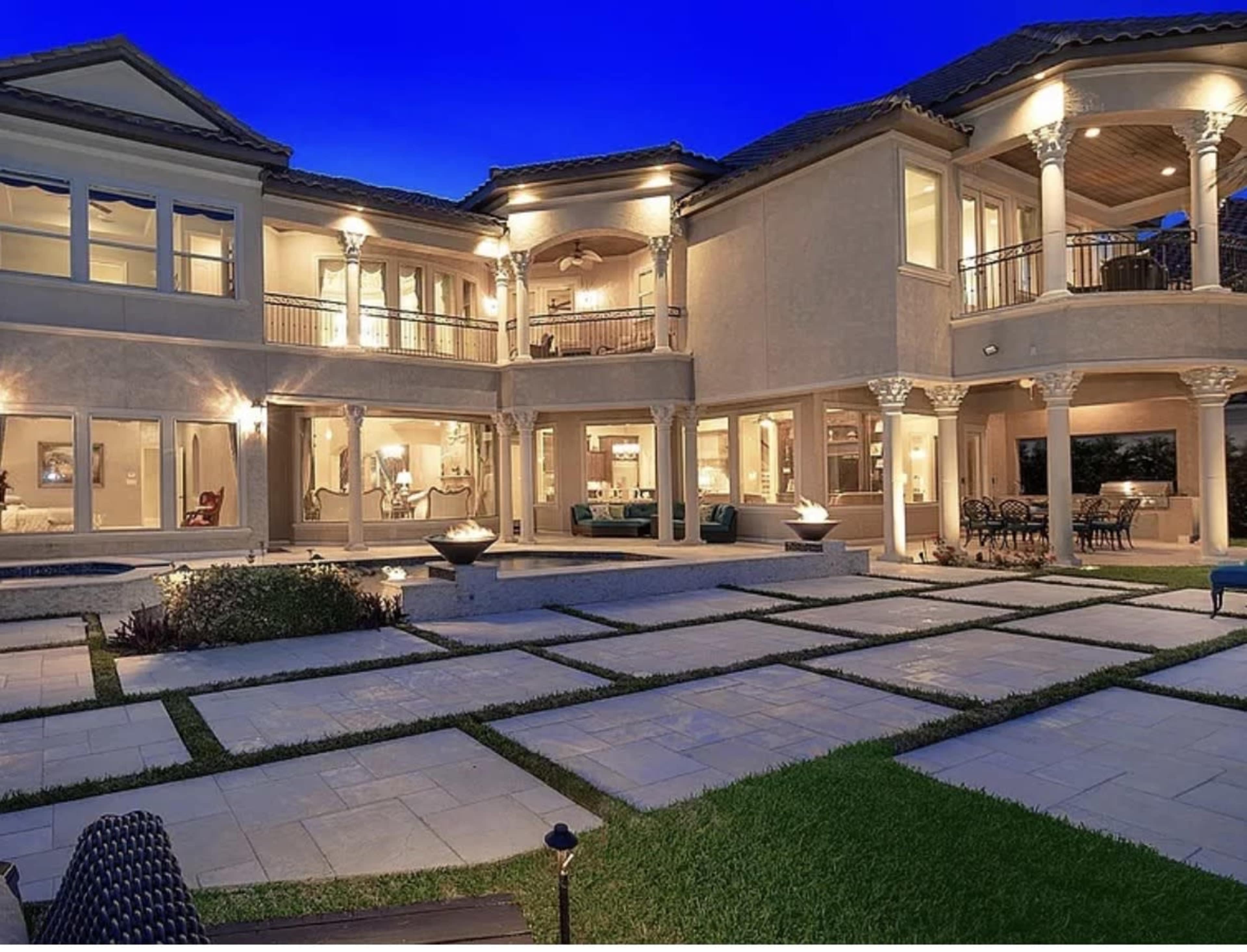 The image shows a large, two-story house with a modern design, surrounded by a neatly landscaped outdoor area featuring square stone paving and illuminated by soft exterior lighting.