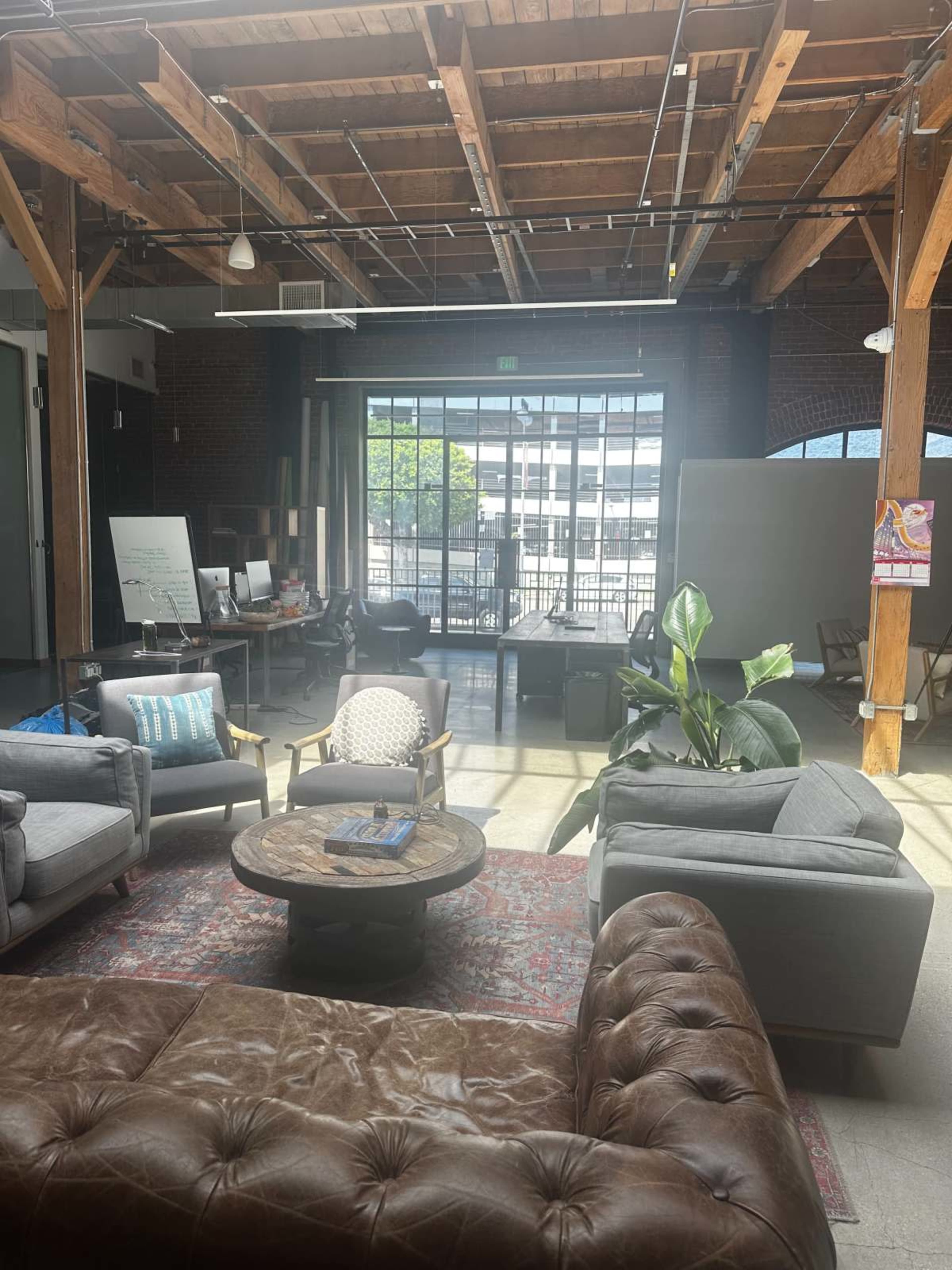 The image shows a spacious office lounge area featuring comfortable seating, a large coffee table, and natural light streaming through large windows.