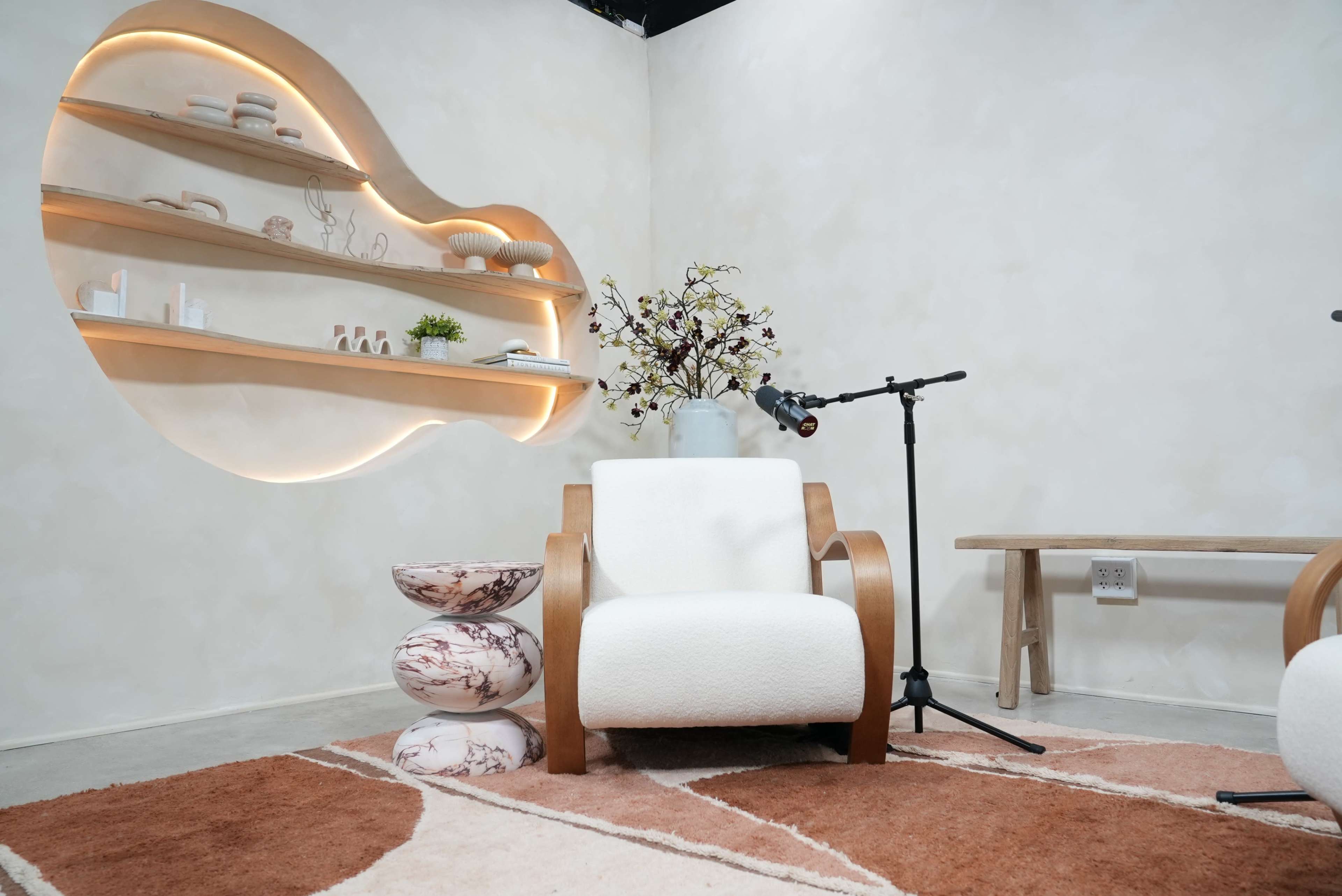 The image shows a modern interior with a cozy white armchair, a microphone on a stand, a decorative shelf with items, and a patterned area rug.