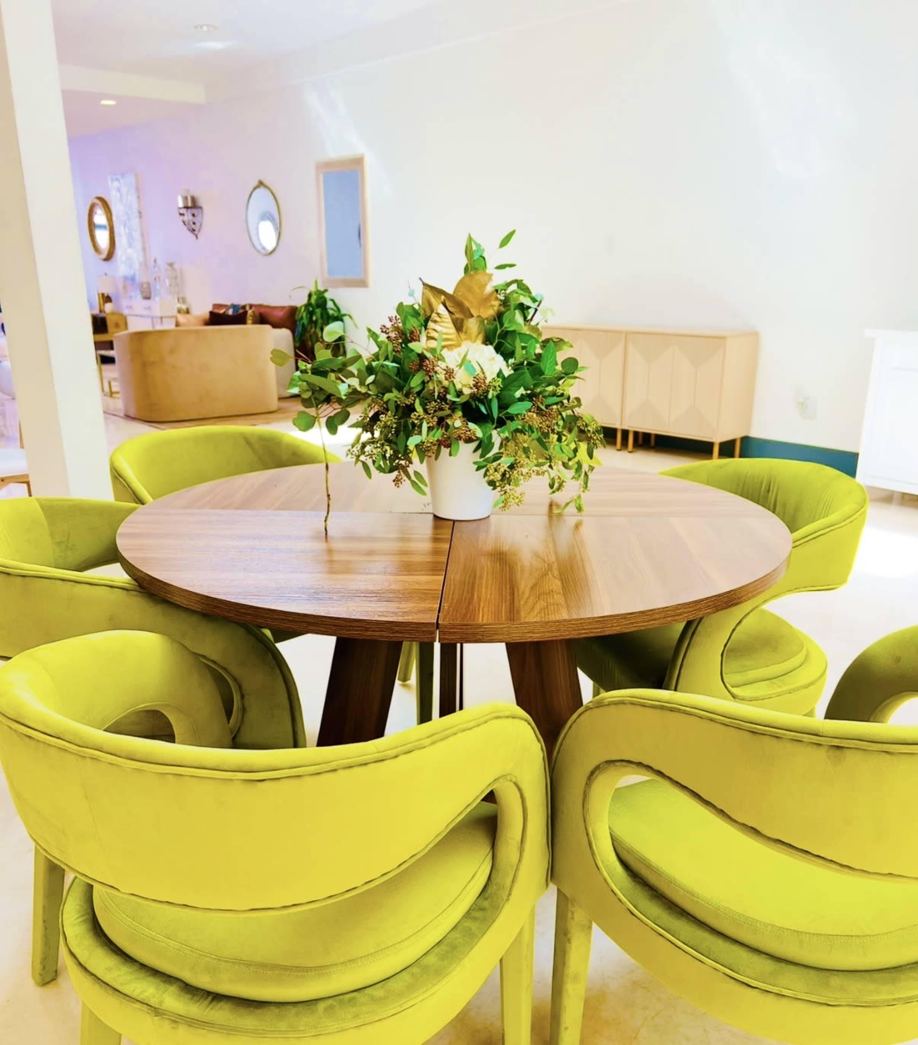 A round wooden dining table with a floral arrangement is surrounded by four green upholstered chairs.