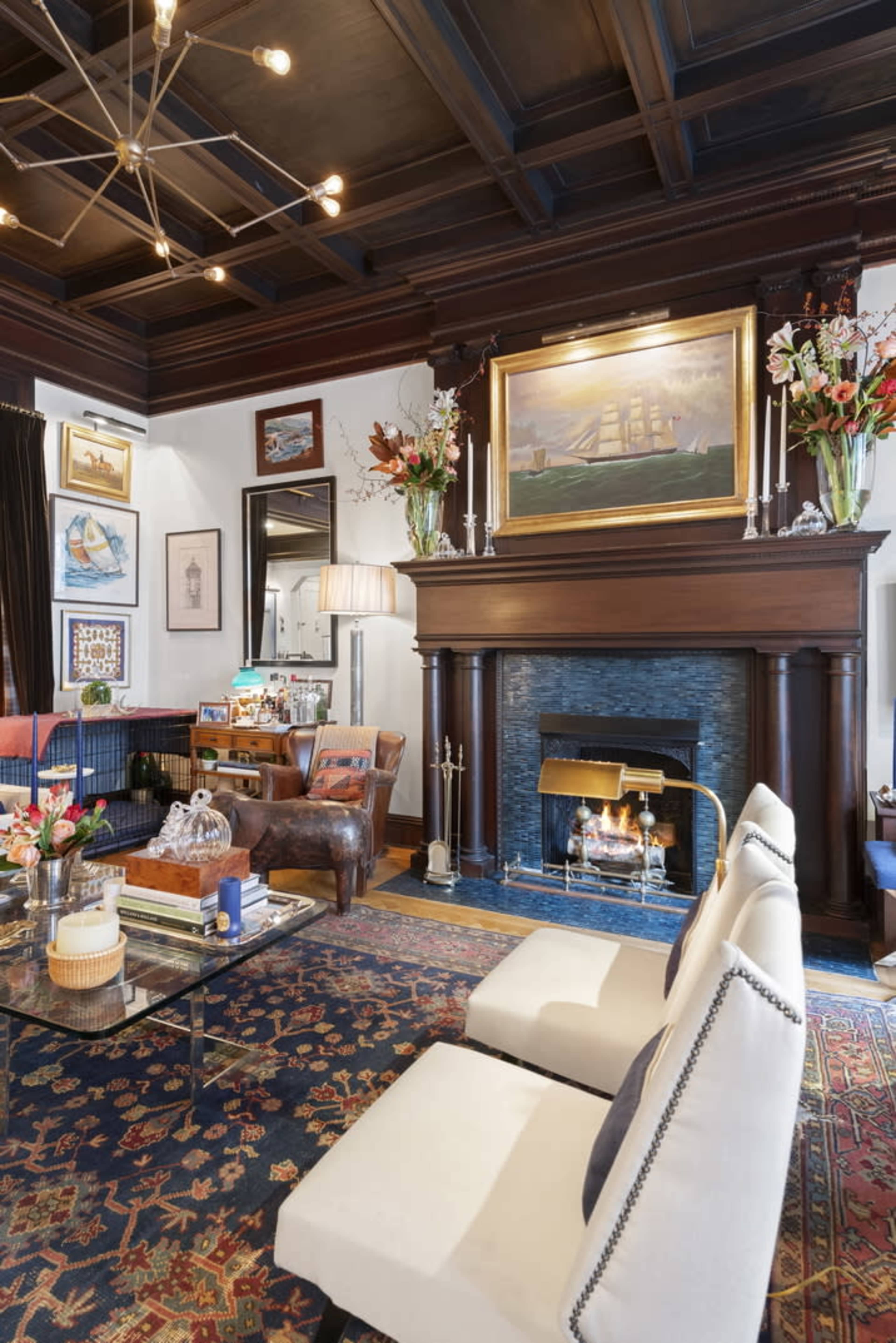 The image depicts a cozy living room featuring a dark wood-paneled ceiling, a large painting above a fireplace, and two upholstered chairs facing a glass coffee table.