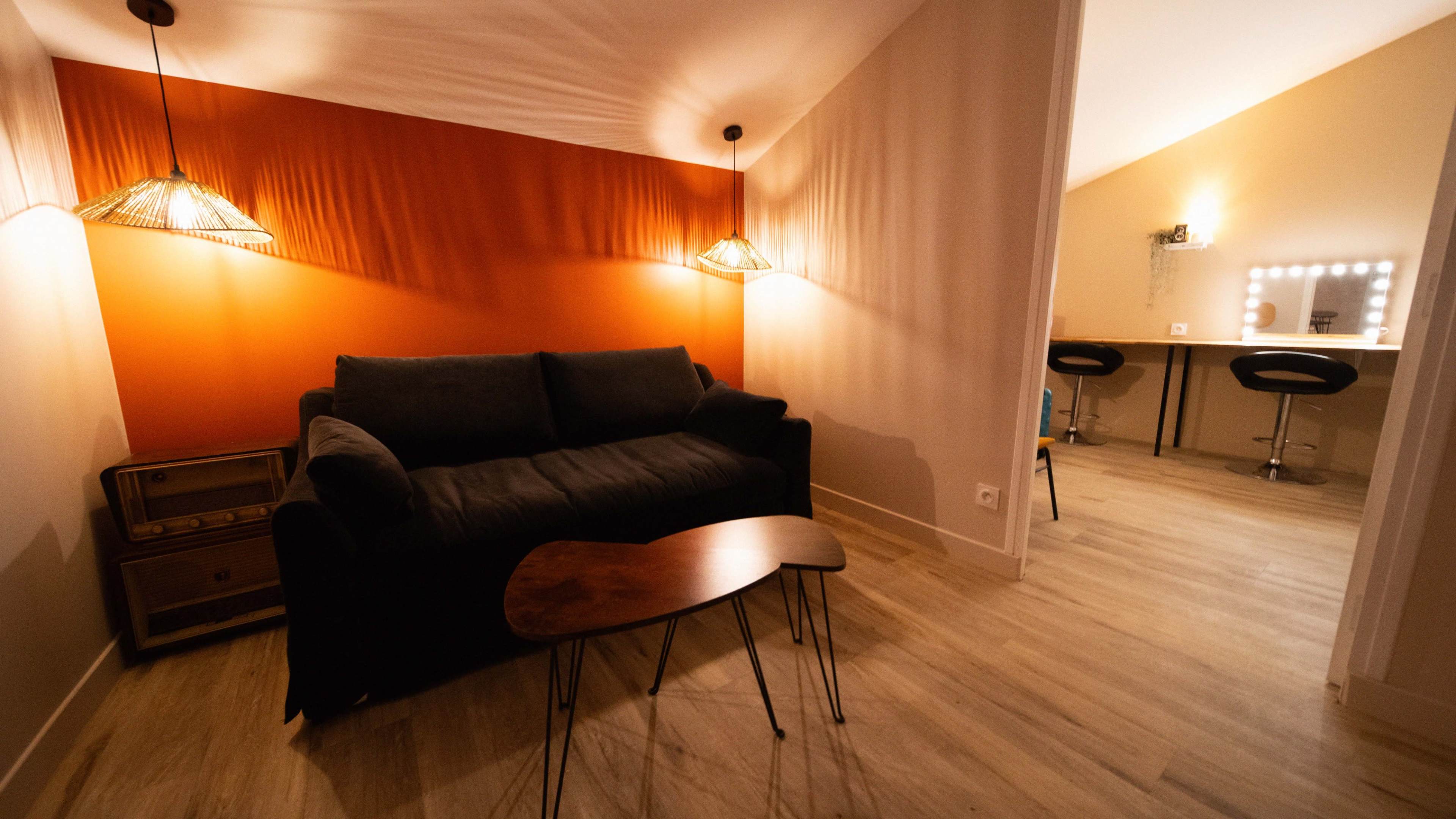 A small living area with a dark sofa, a wooden coffee table, and a lighted vanity area in the background against a partially orange wall.