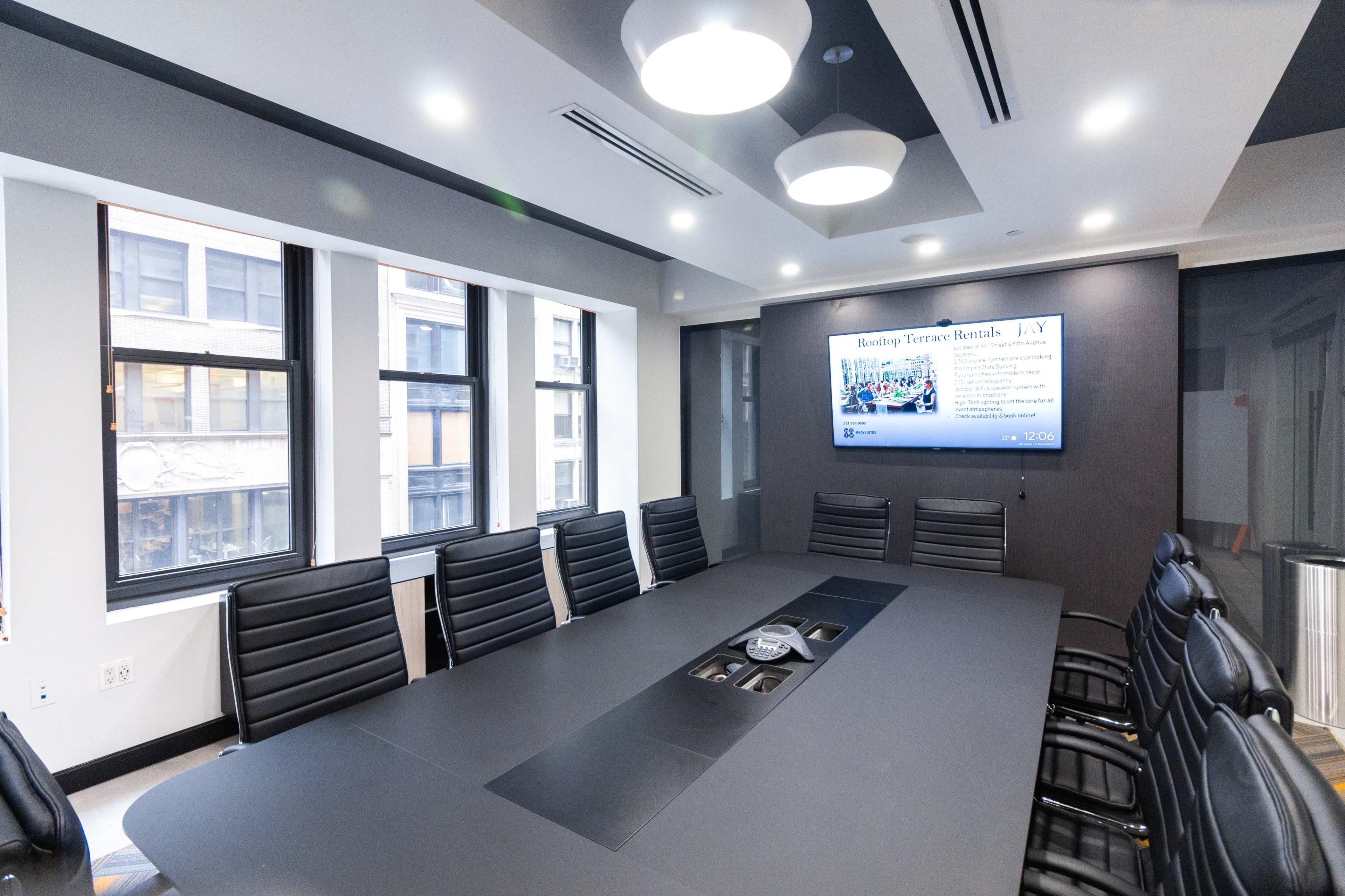 A modern conference room features a large rectangular table surrounded by black leather chairs, with a screen displaying information on one wall and windows letting in natural light.