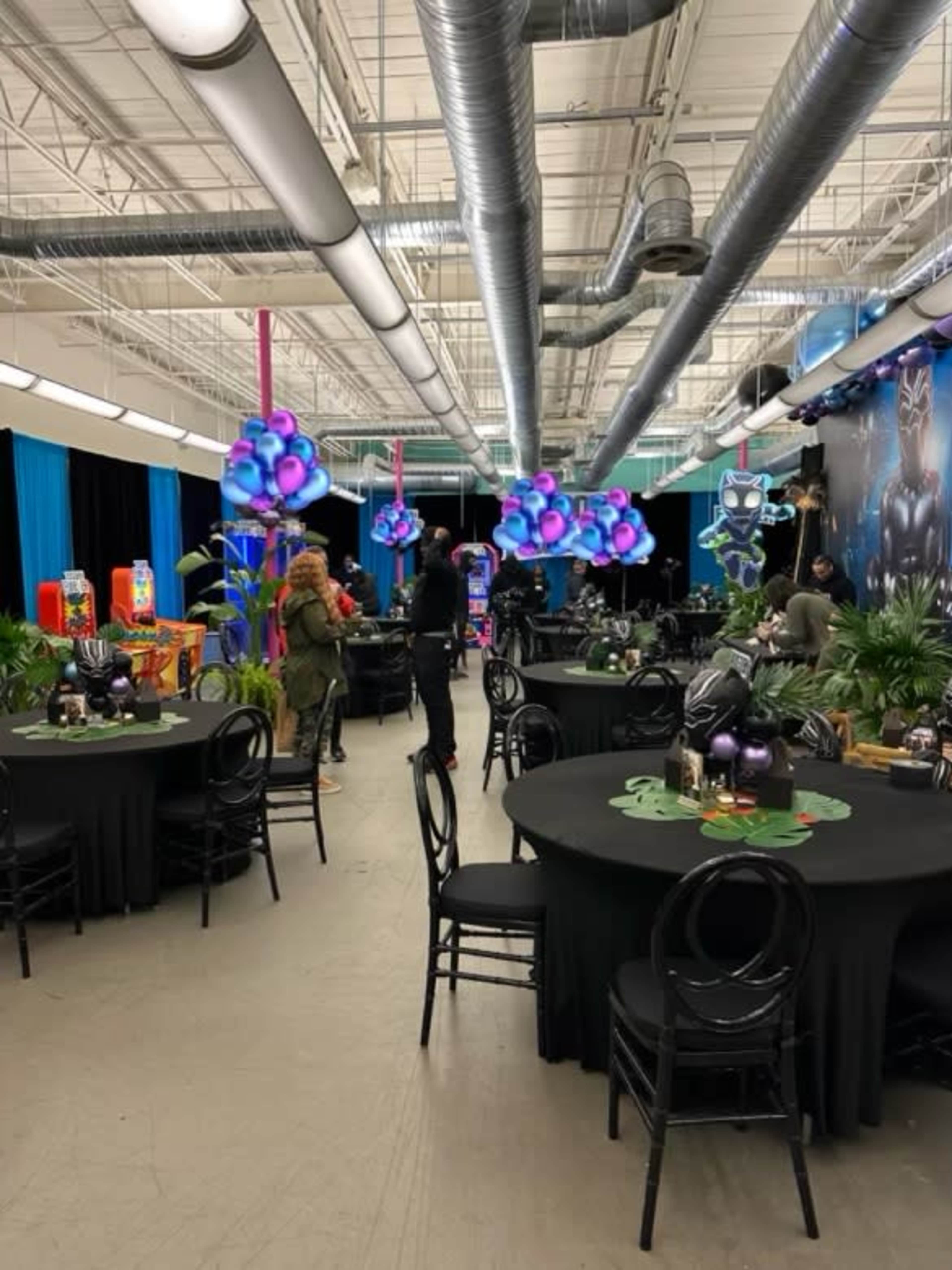 A spacious indoor area set up for an event, featuring round black tables, decorative plants, and colorful hanging light fixtures.