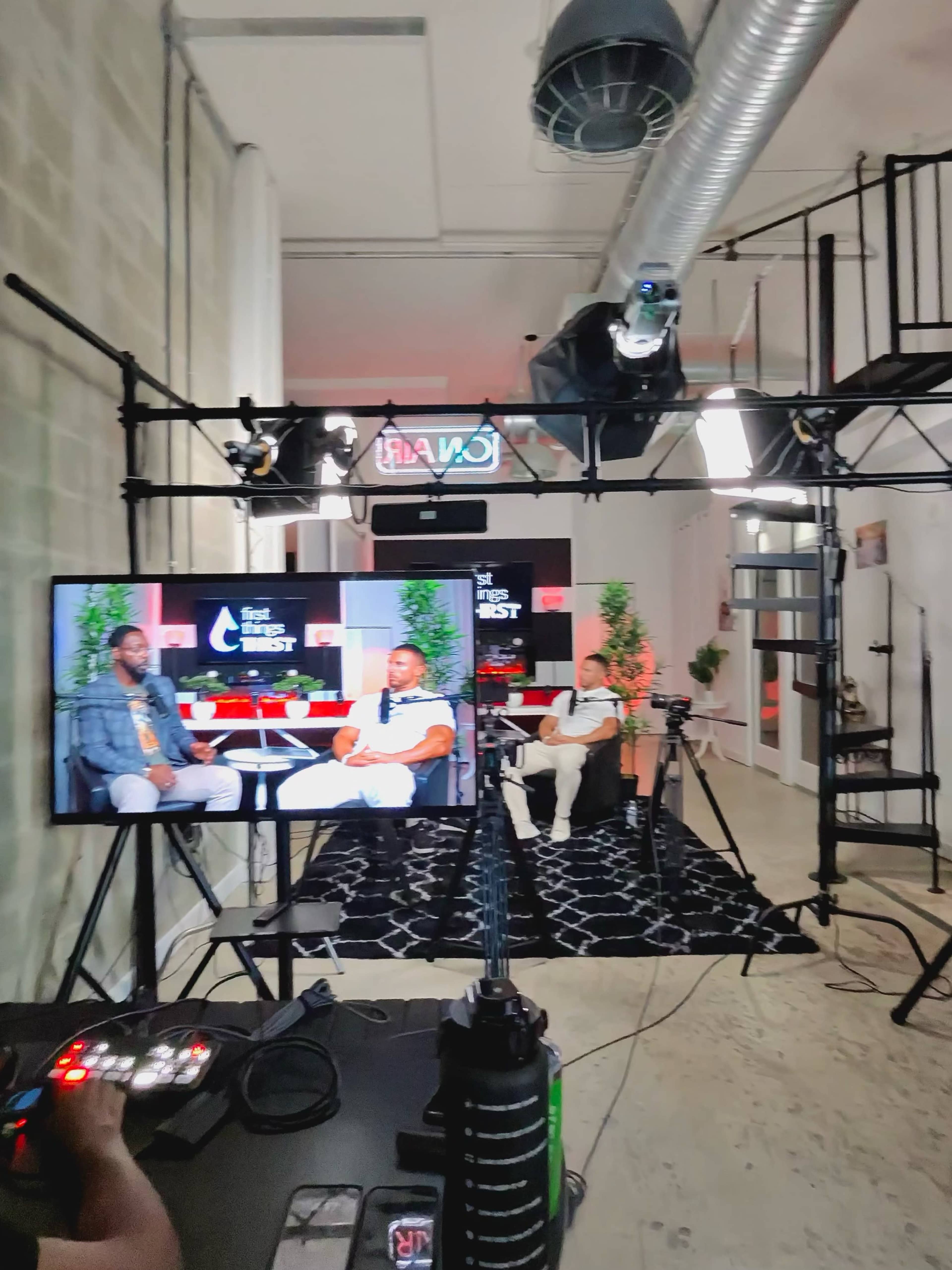 The image shows a studio setup for a video interview, featuring two seated speakers, cameras, and lighting equipment, with a large screen displaying graphics in the background.
