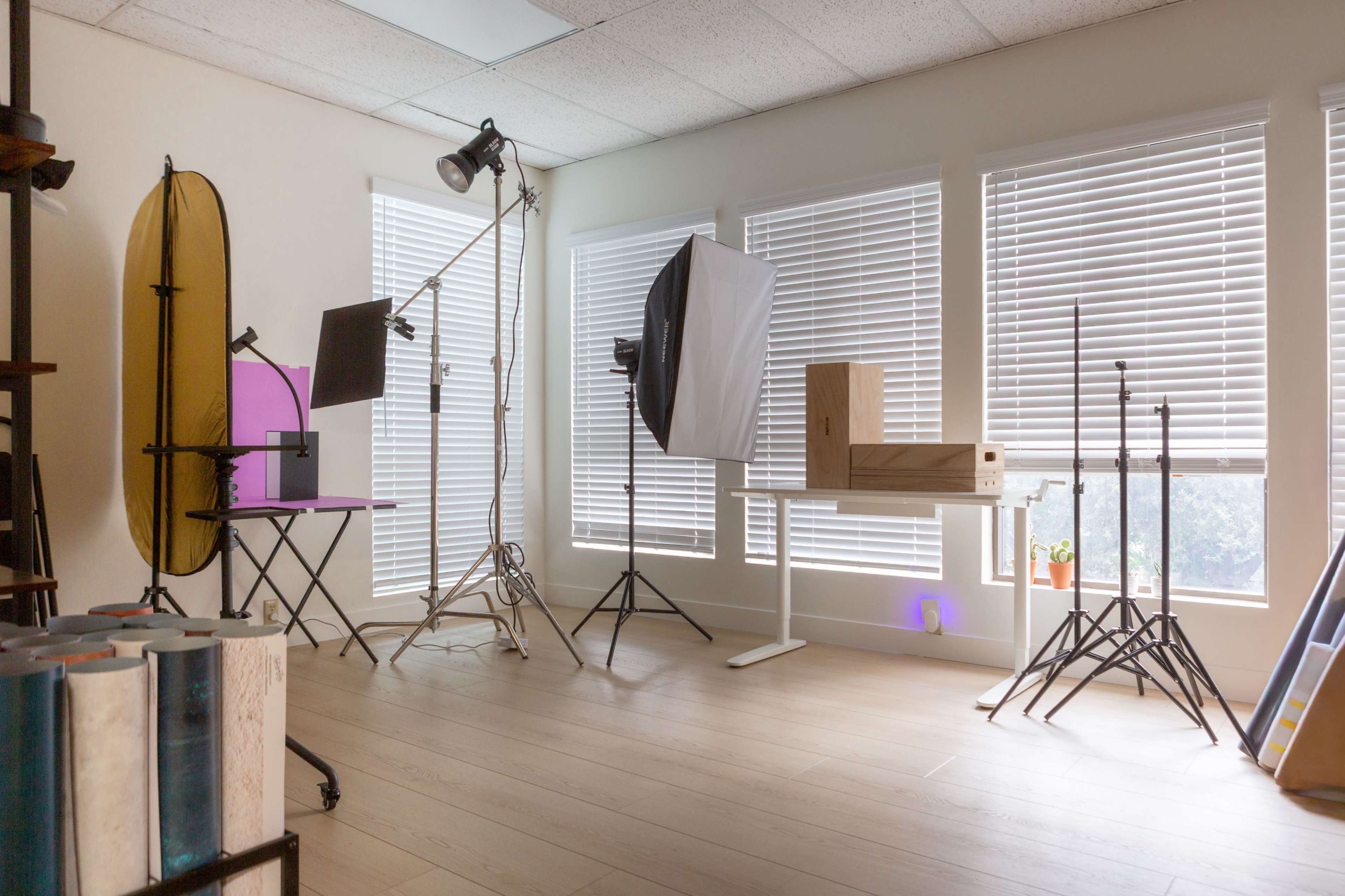 The image shows a well-lit photography studio with various lighting equipment, a table, and stands arranged near large windows with blinds.