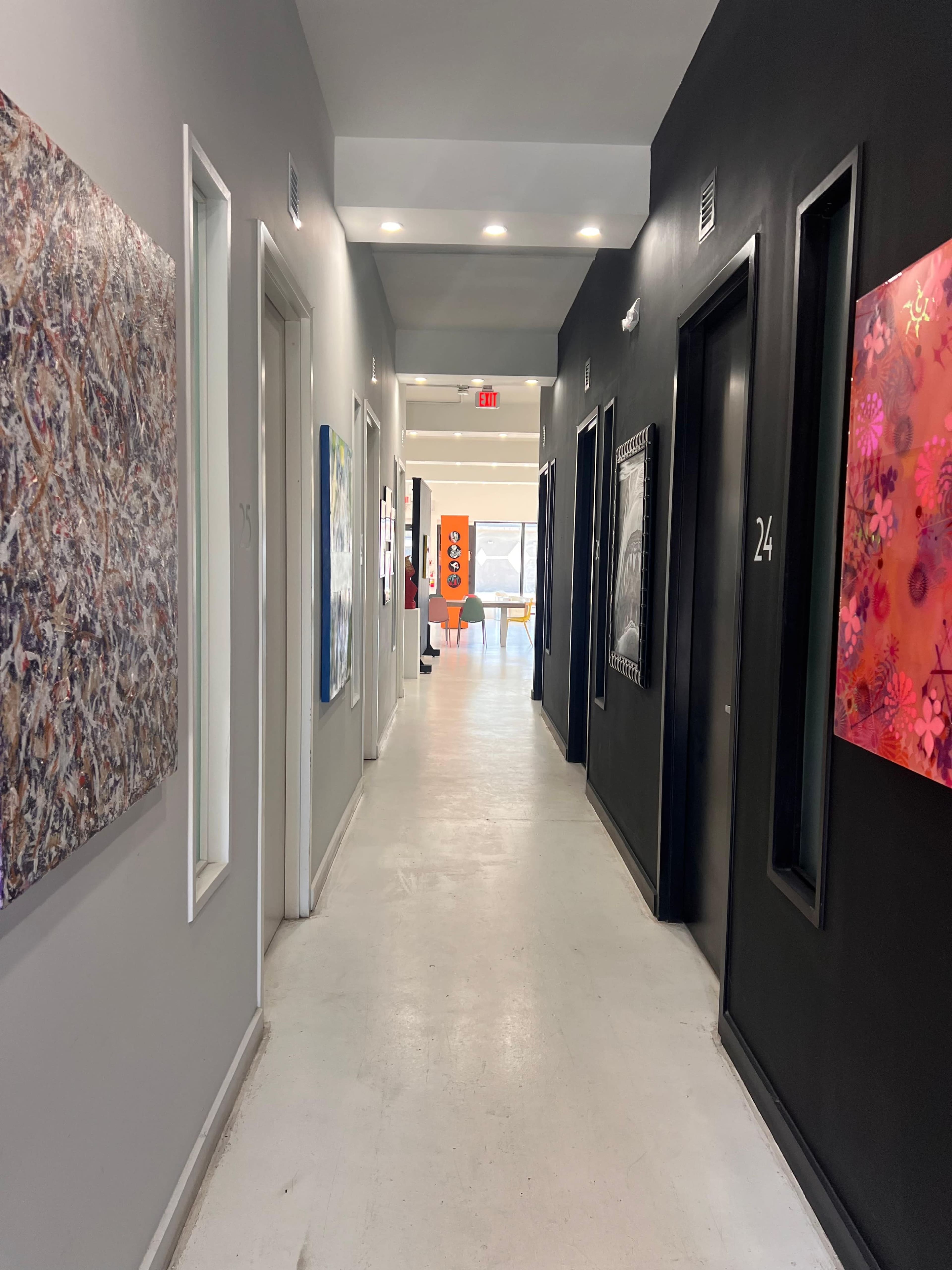 A long corridor features art on the walls, with doors numbered along one side and natural light illuminating the space from windows at the far end.