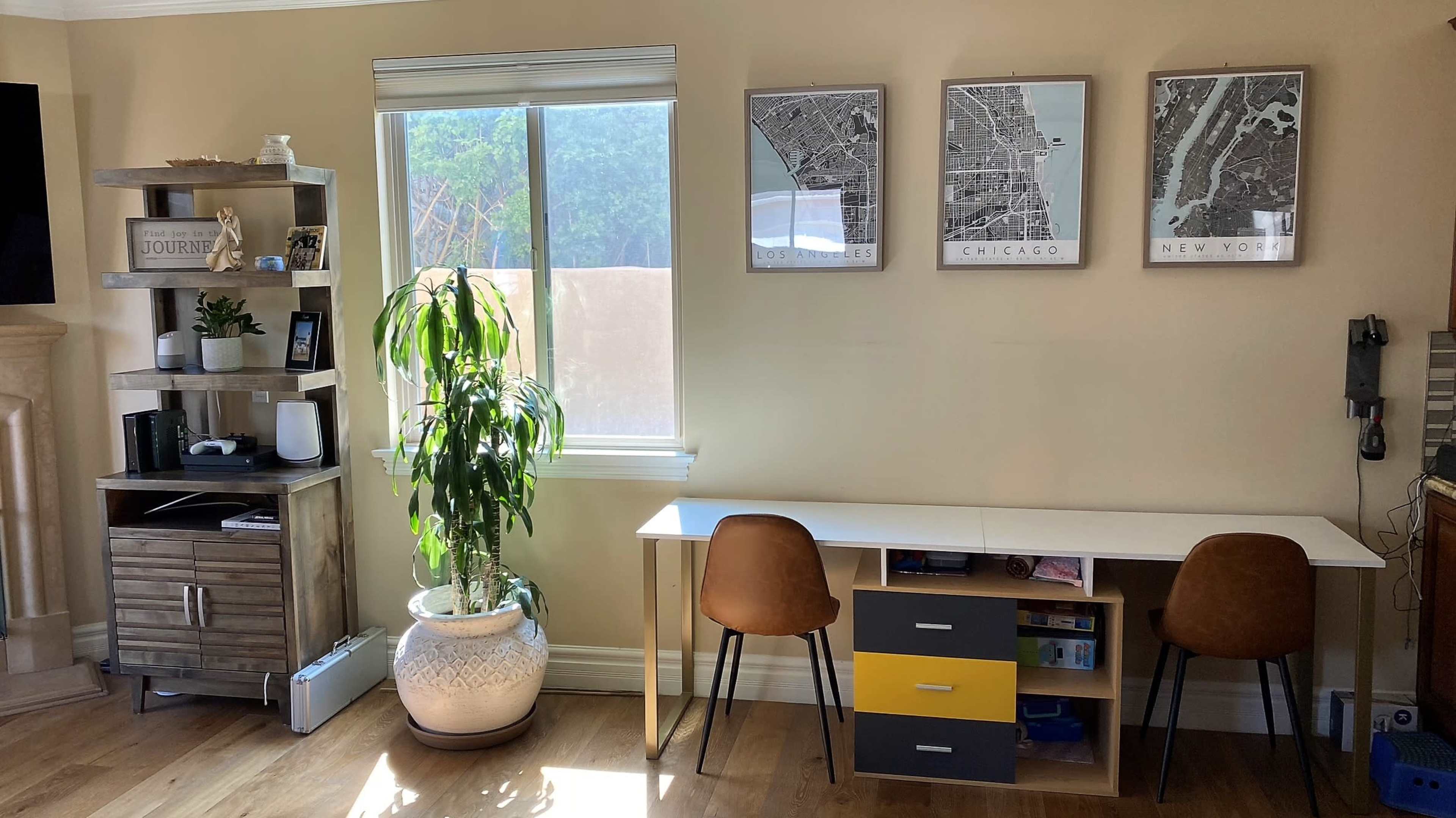A bright room with a window, a desk with two chairs, and a plant next to a shelf displaying decor and maps of various cities.